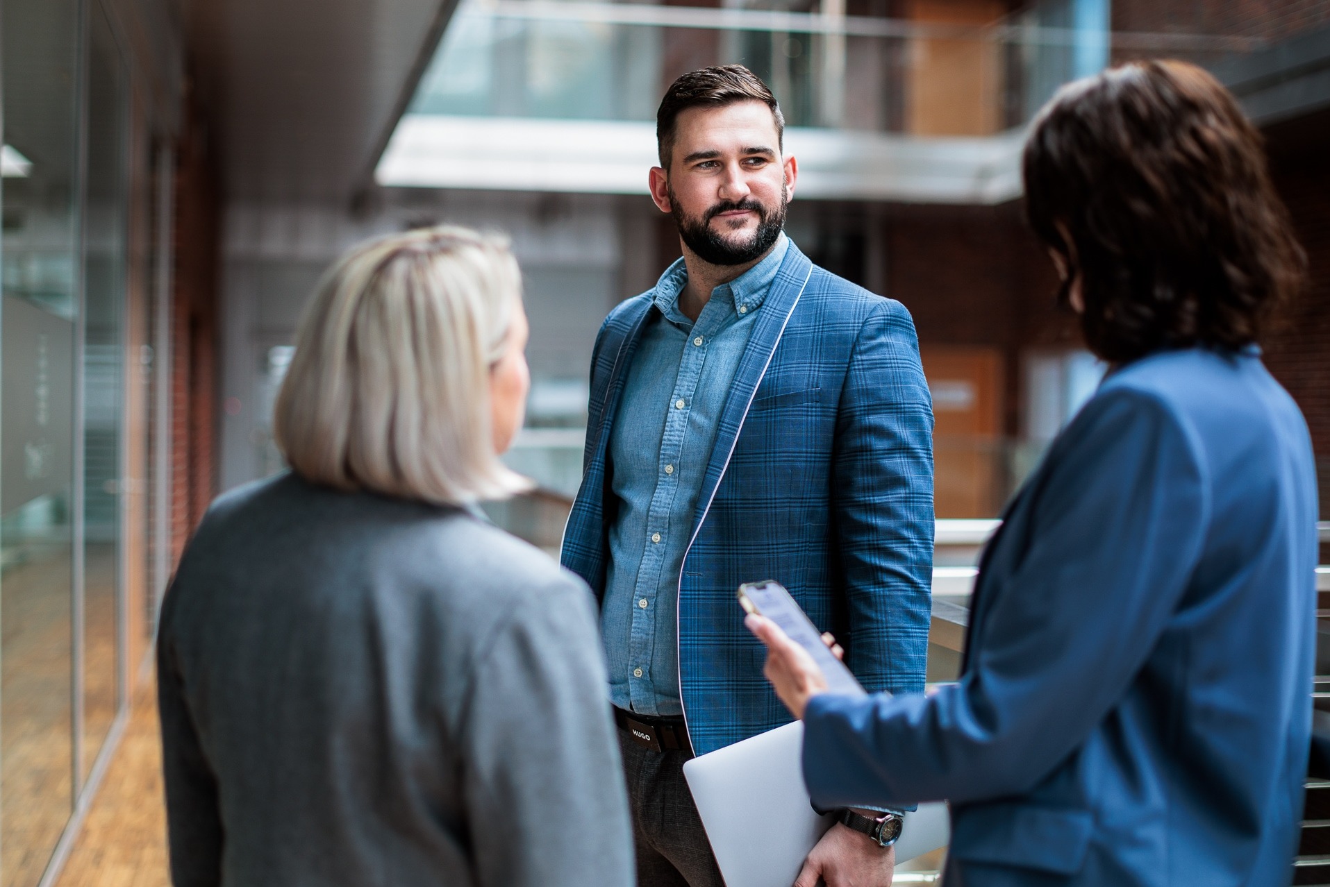 Spotkanie biznesowe w nowoczesnym biurze: mężczyzna w niebieskiej marynarce rozmawia z dwiema kobietami, jedna z laptopem i telefonem. Jasne wnętrze, profesjonalna atmosfera.