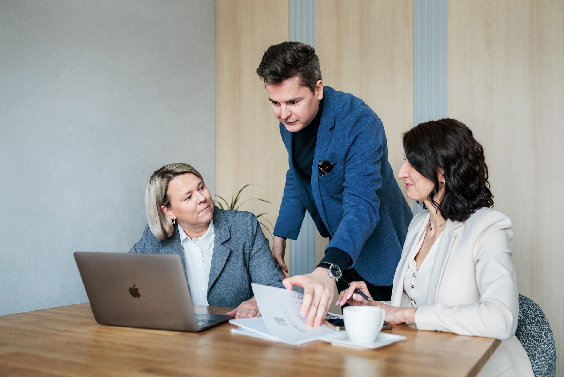 Spotkanie biznesowe: mężczyzna w niebieskiej marynarce omawia dokumenty z dwiema kobietami przy biurku z laptopem i filiżanką kawy.