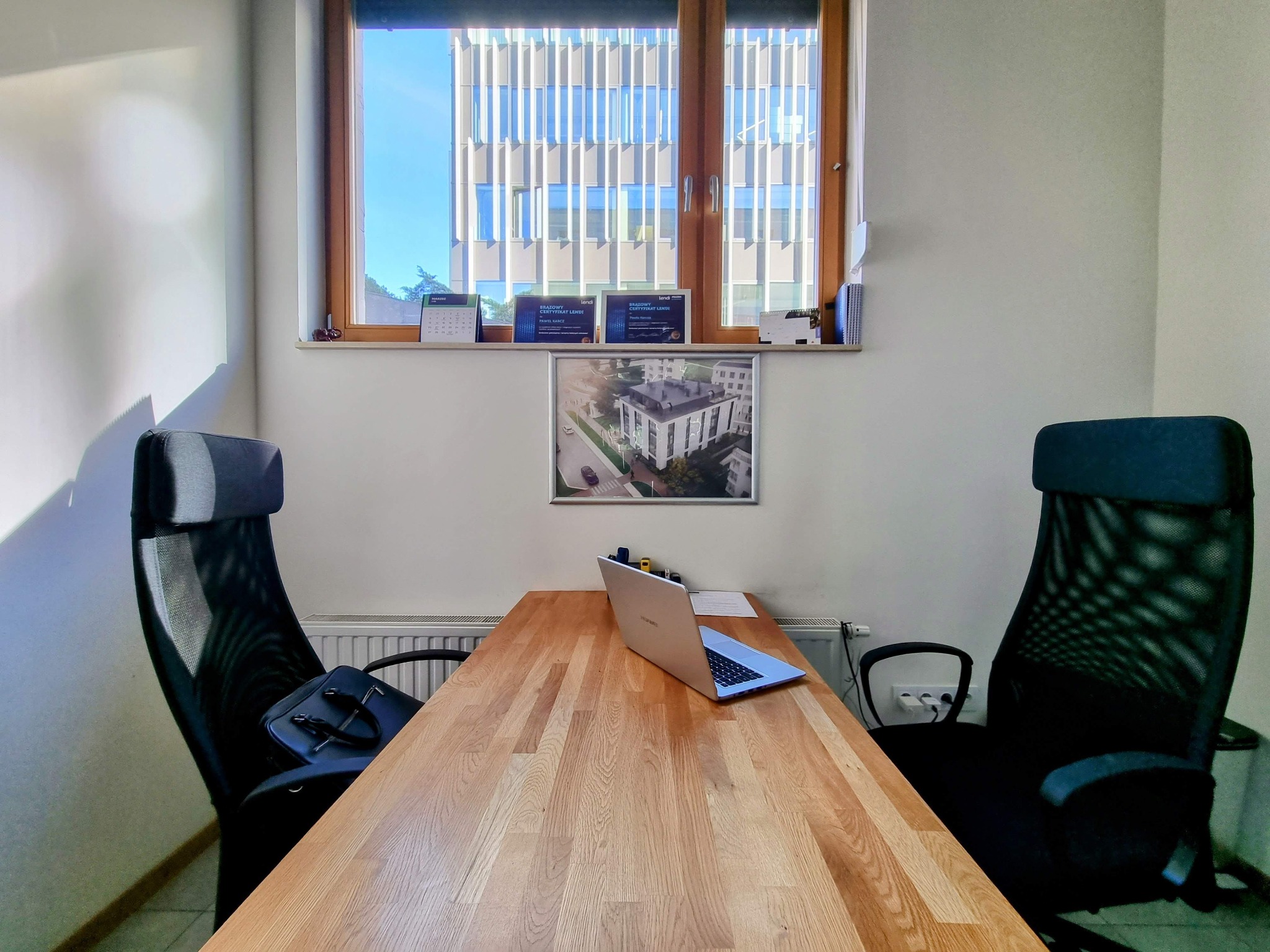 Biuro doradcy w Krakowie: drewniany stół, laptop, dwa krzesła biurowe, okno z widokiem na nowoczesny budynek i certyfikaty na parapecie.