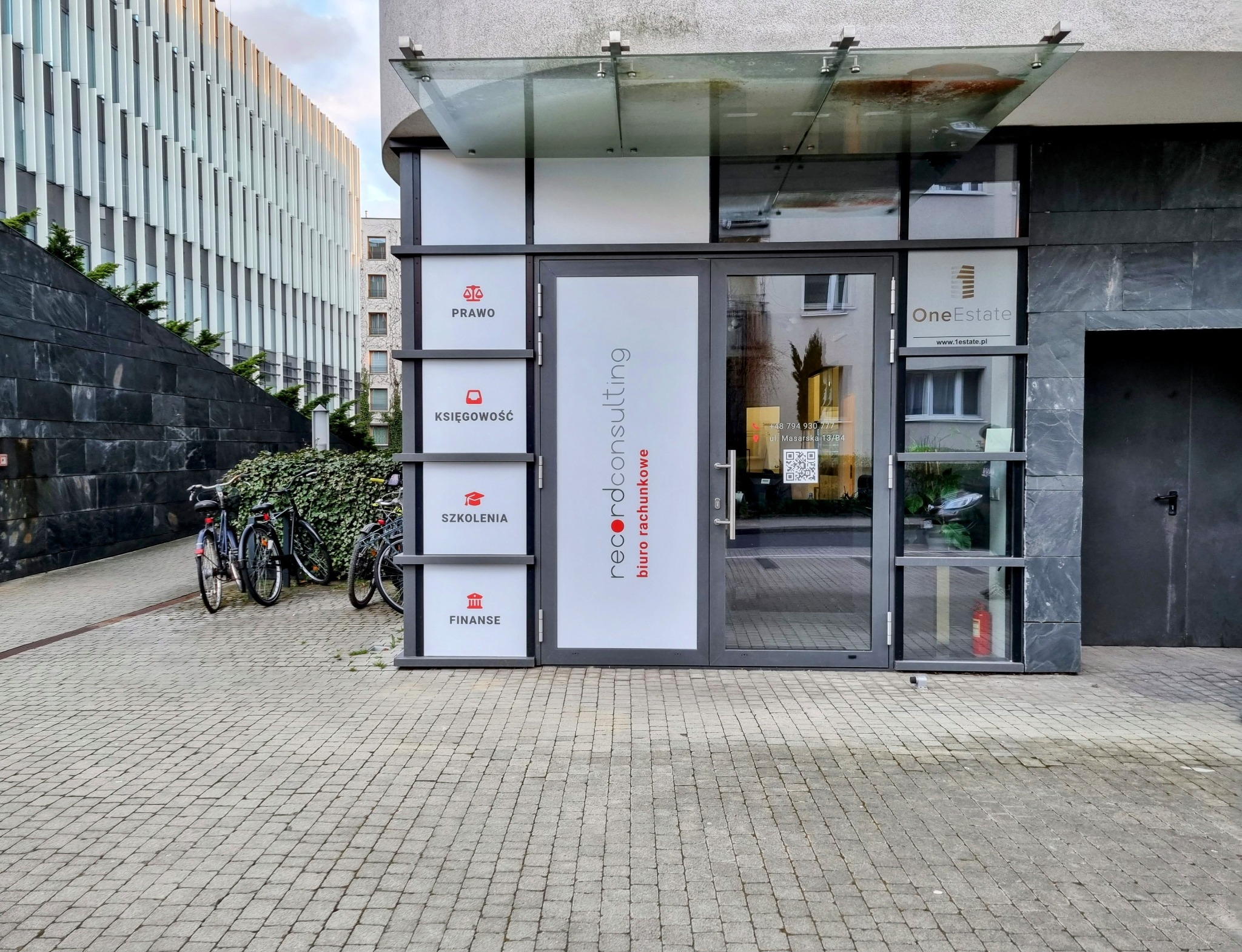 Wejście do biura Record Consulting z informacjami o świadczonych usługach: prawo, księgowość, szkolenia, finanse. Obok wejścia szyld One Estate. Na chodniku zaparkowane rowery.