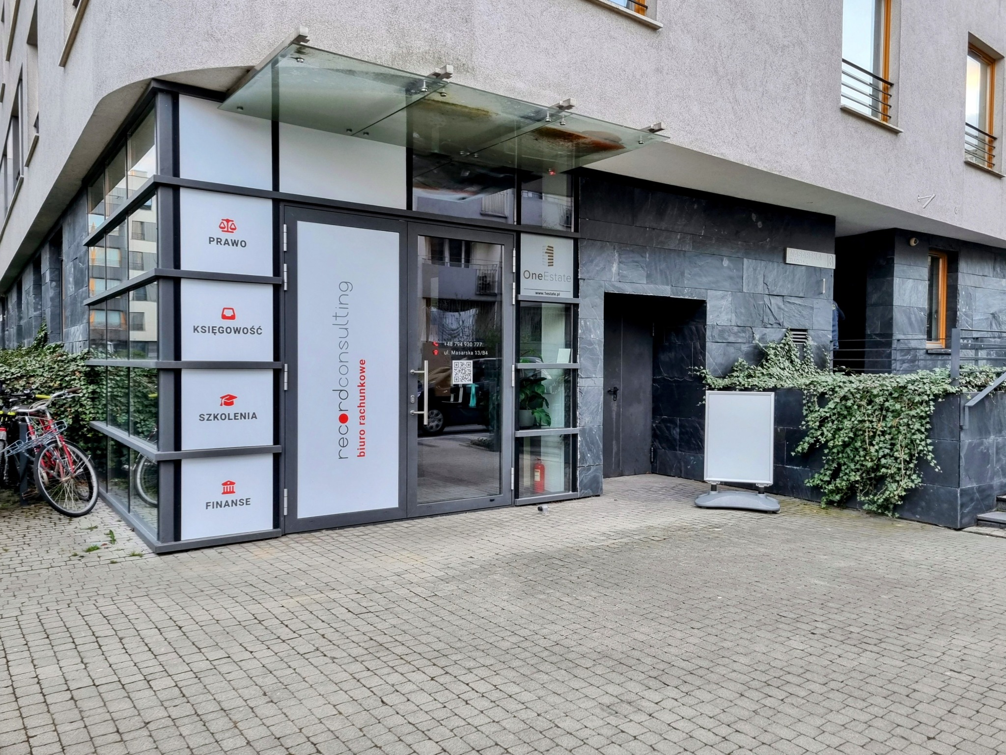 Wejście do biura rachunkowego Record Consulting z informacjami o usługach: prawo, księgowość, szkolenia i finanse. Widoczny rower zaparkowany obok.