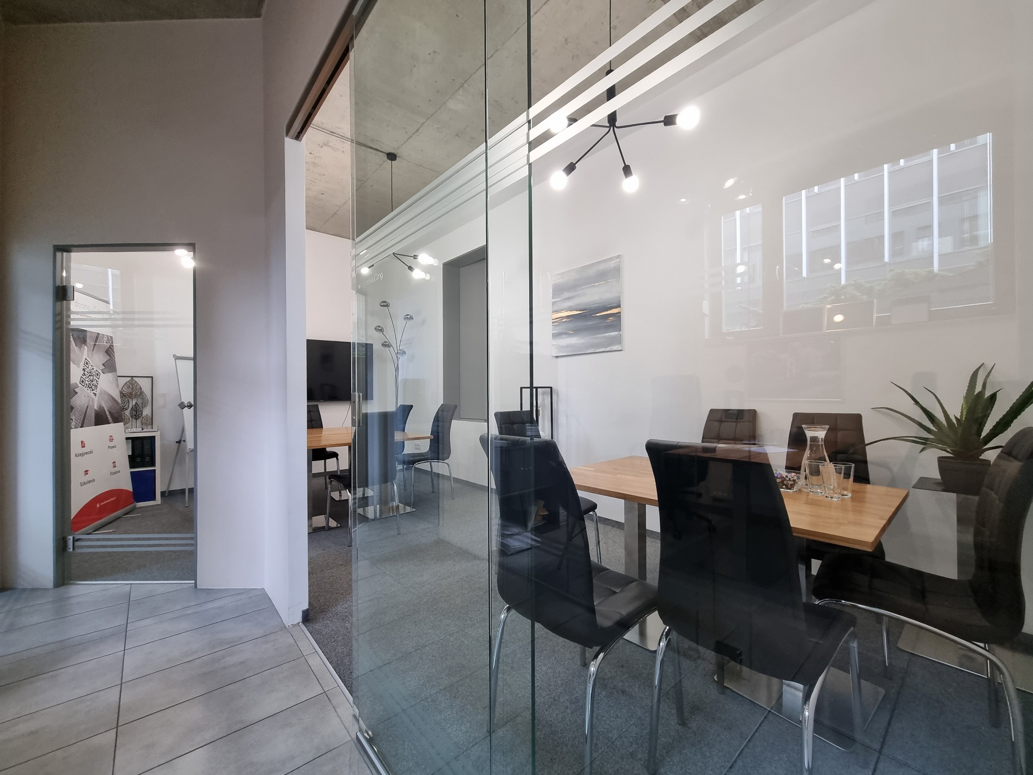 Nowoczesne biuro z szklanymi ścianami, widoczna sala konferencyjna z drewnianym stołem, czarnymi krzesłami, rośliną doniczkową i minimalistyczną lampą sufitową.