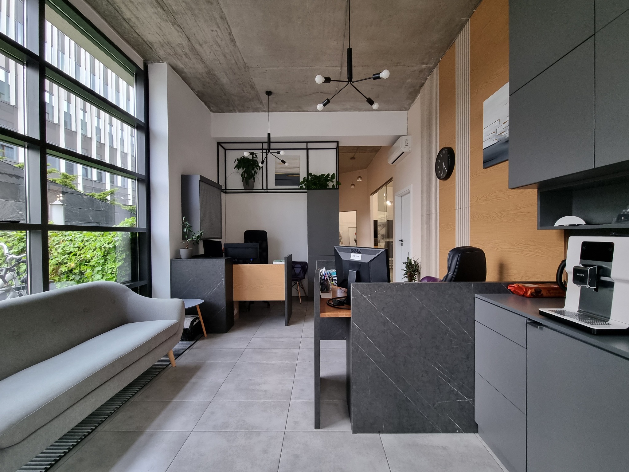 Nowoczesne biuro doradcy finansowego z szarą sofą, biurkami z marmurowym wykończeniem i dużym oknem z widokiem na zieleń.