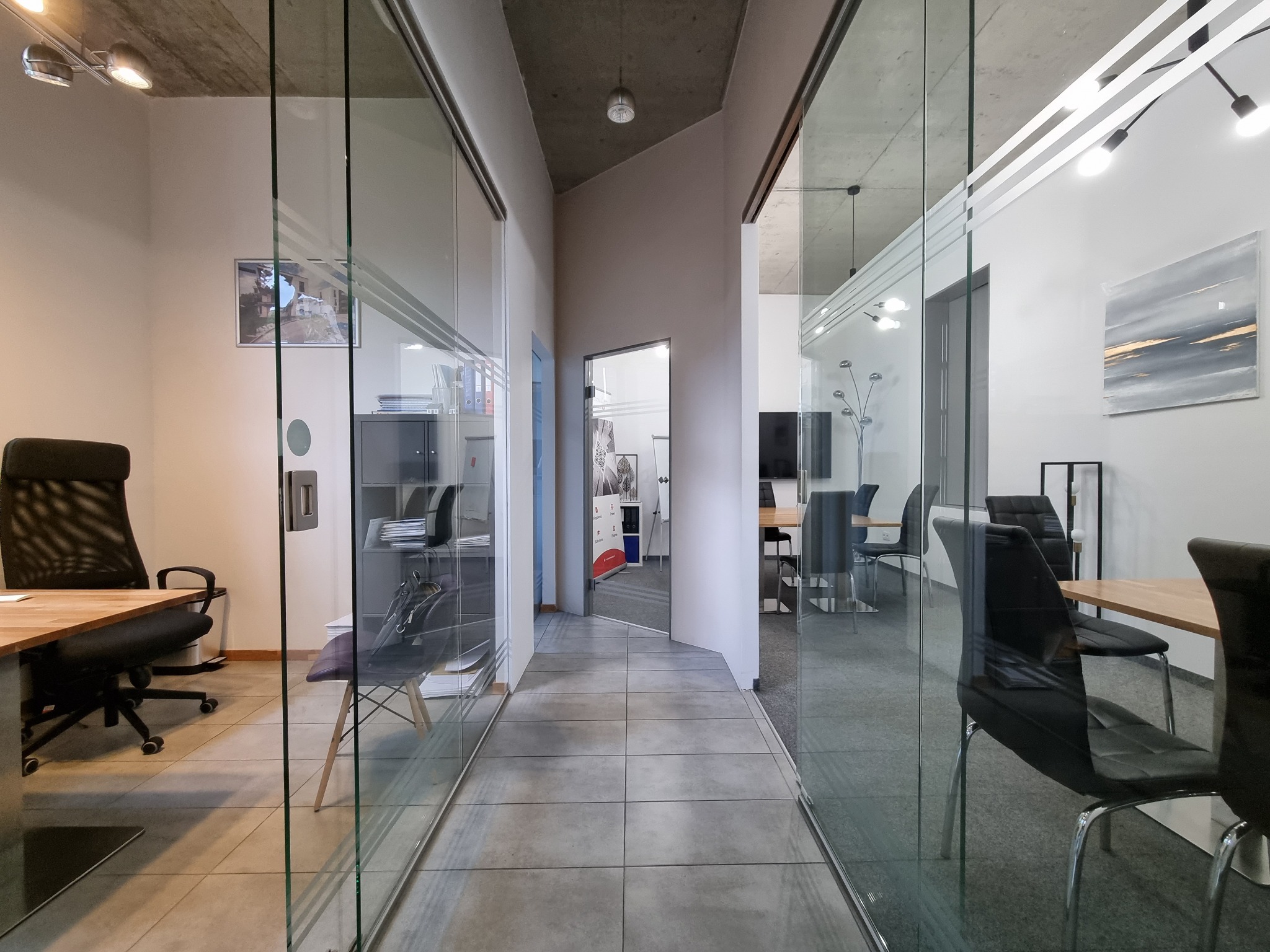 Wnętrze nowoczesnego biura z przeszklonymi ścianami, widoczne biurka, krzesła i oświetlenie sufitowe.