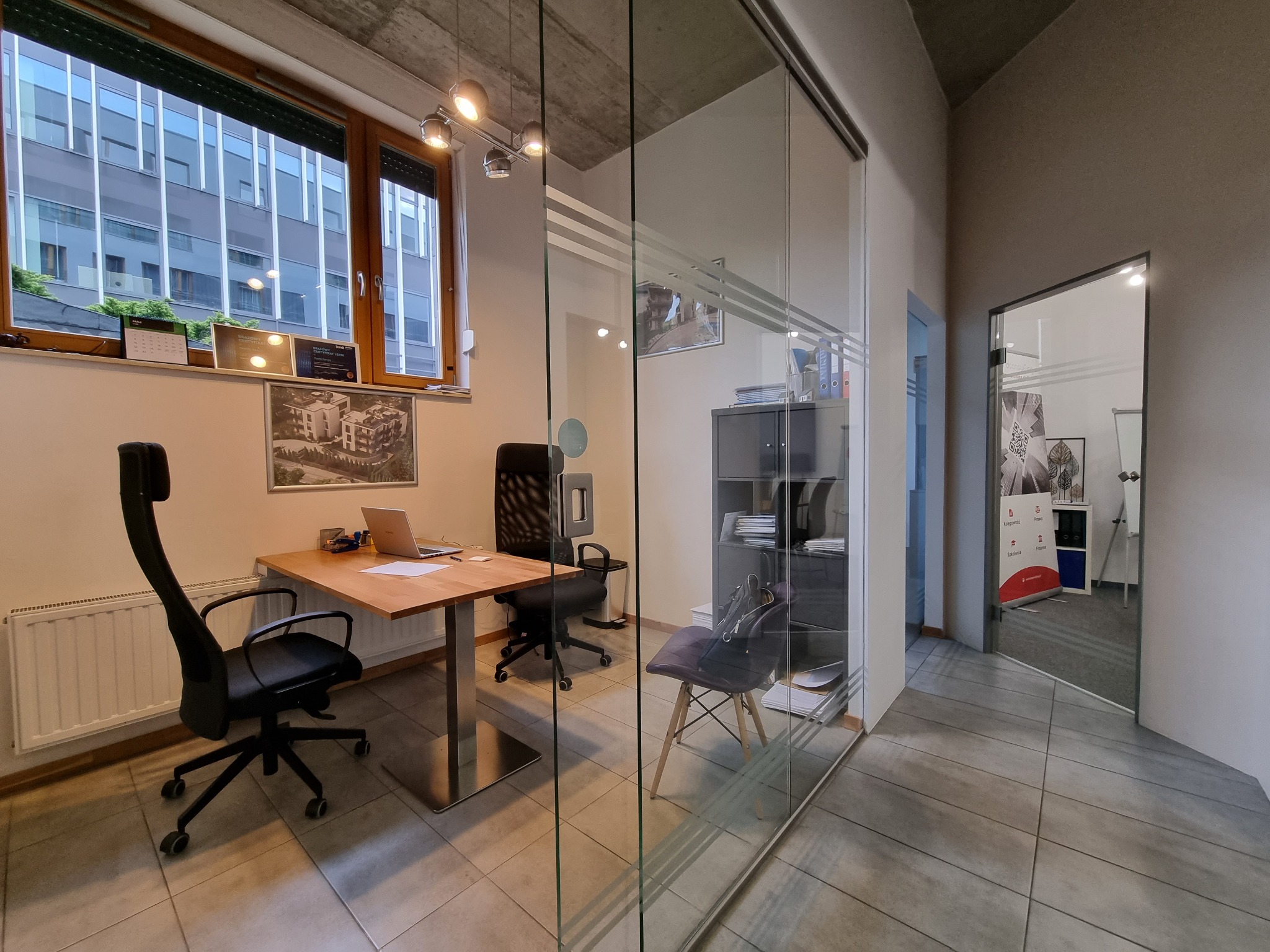 Nowoczesne biuro z przeszklonymi ścianami, biurkiem z laptopem, czarnymi krzesłami obrotowymi i widokiem na budynek zza okna. Na parapecie kalendarz i certyfikaty.