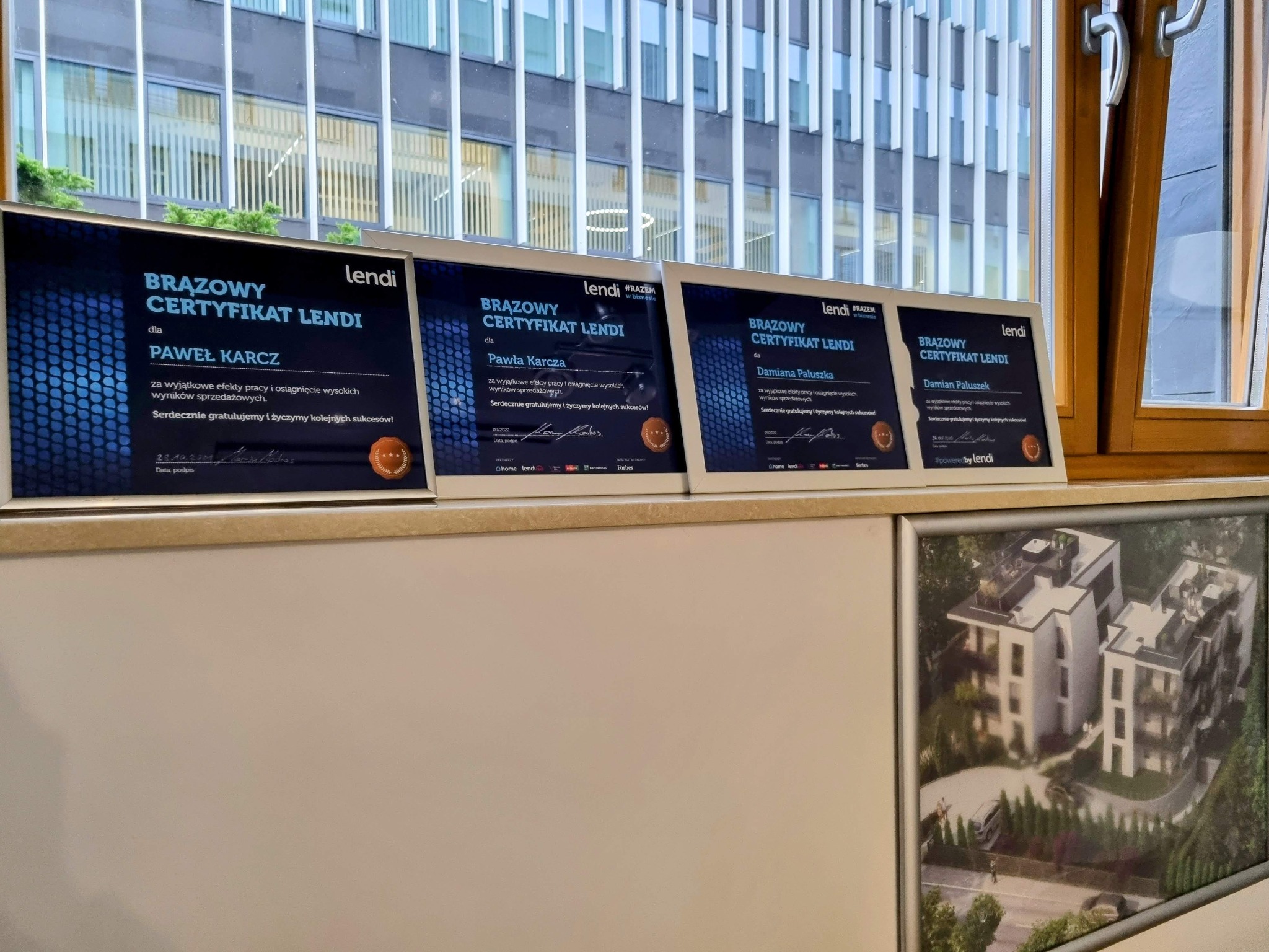 Cztery oprawione brązowe certyfikaty Lendi, z nazwami Pawła Karcza i Damiana Paluszka, zawieszone na ścianie z widokiem na budynek biurowy i osiedle mieszkaniowe.