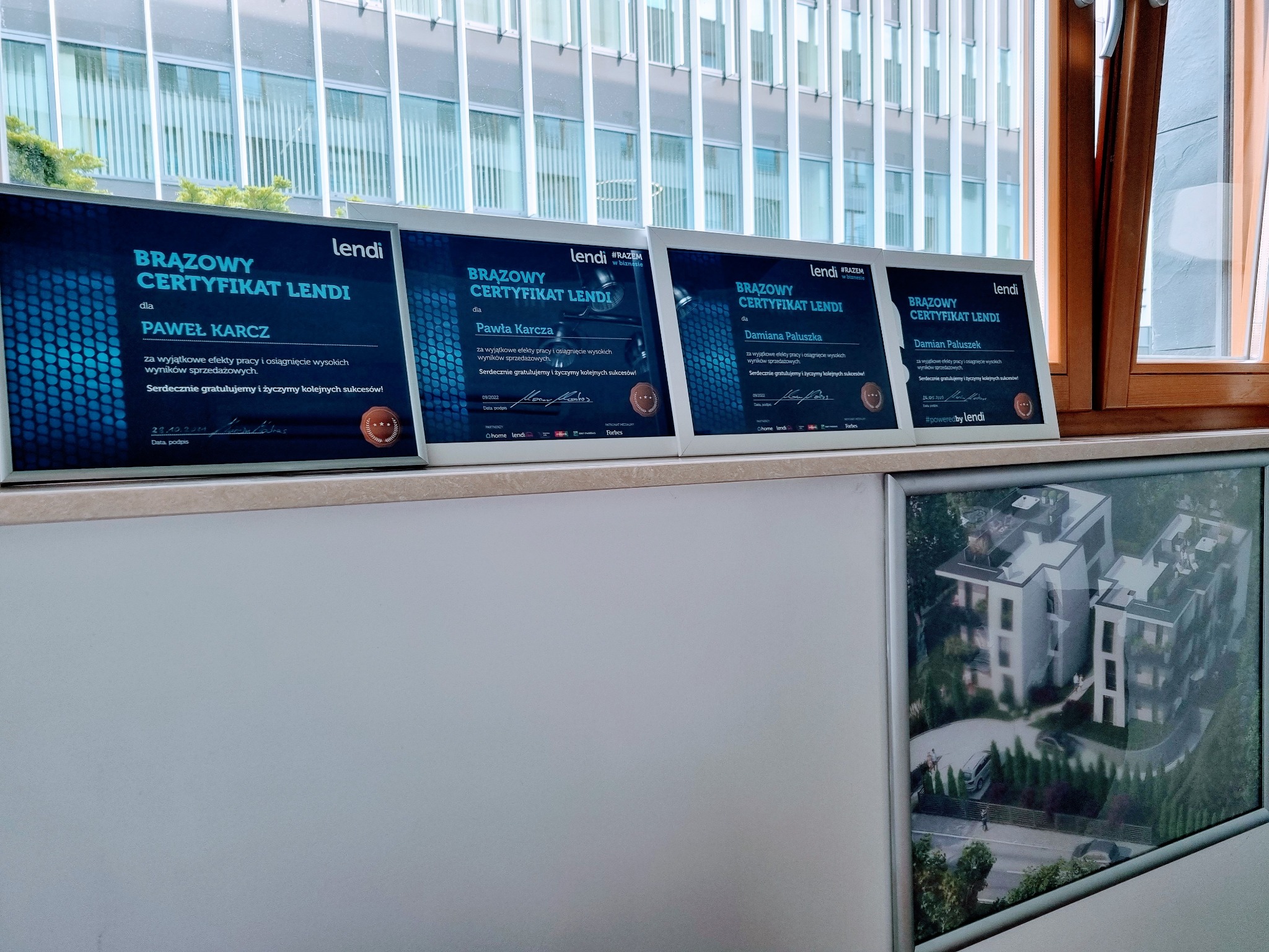 Cztery oprawione brązowe certyfikaty Lendi na blacie z widokiem na budynek biurowy, obok ramka ze zdjęciem osiedla mieszkaniowego.