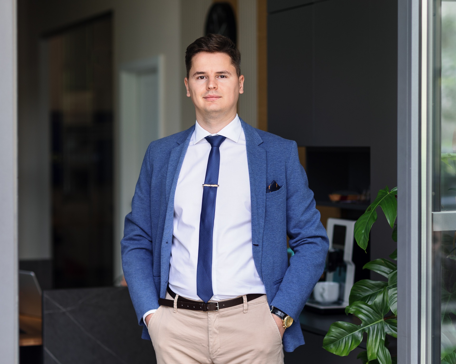 Młody mężczyzna w błękitnej marynarce, białej koszuli i granatowym krawacie ze spinką, stoi z rękami w kieszeniach beżowych spodni, w tle widoczne wnętrze nowoczesnego biura z roślinami doniczkowymi.
