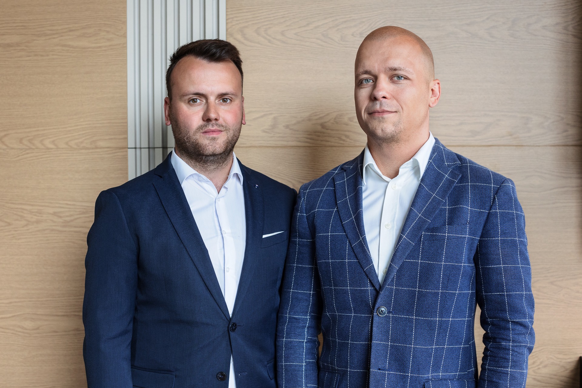 Portret dwóch mężczyzn w garniturach na tle drewnianej ściany, jeden w gładkim granatowym garniturze, drugi w garniturze w niebieską kratę.