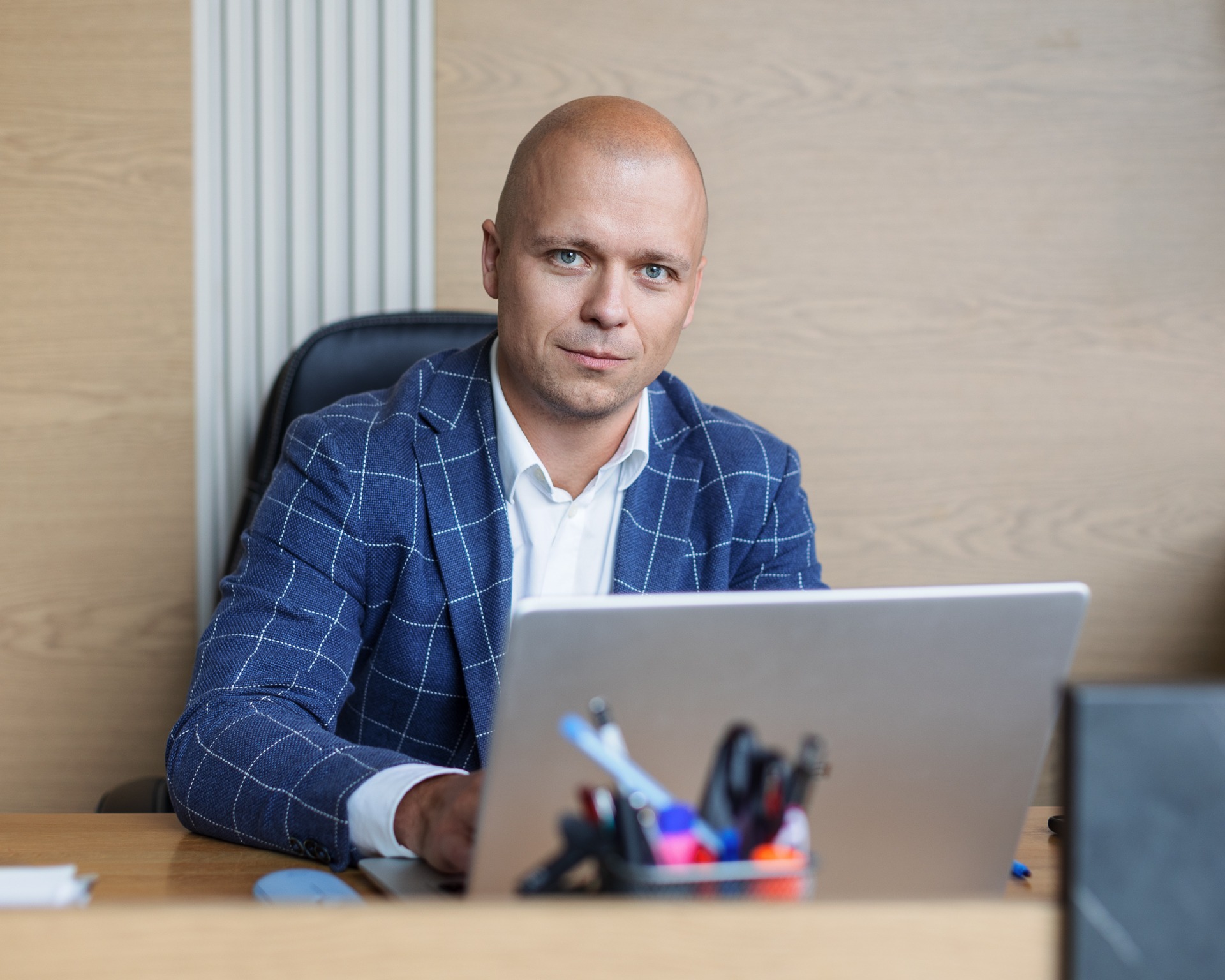 Mężczyzna w niebieskiej marynarce w kratę siedzi za biurkiem z laptopem, spoglądając w obiektyw z lekkim uśmiechem.