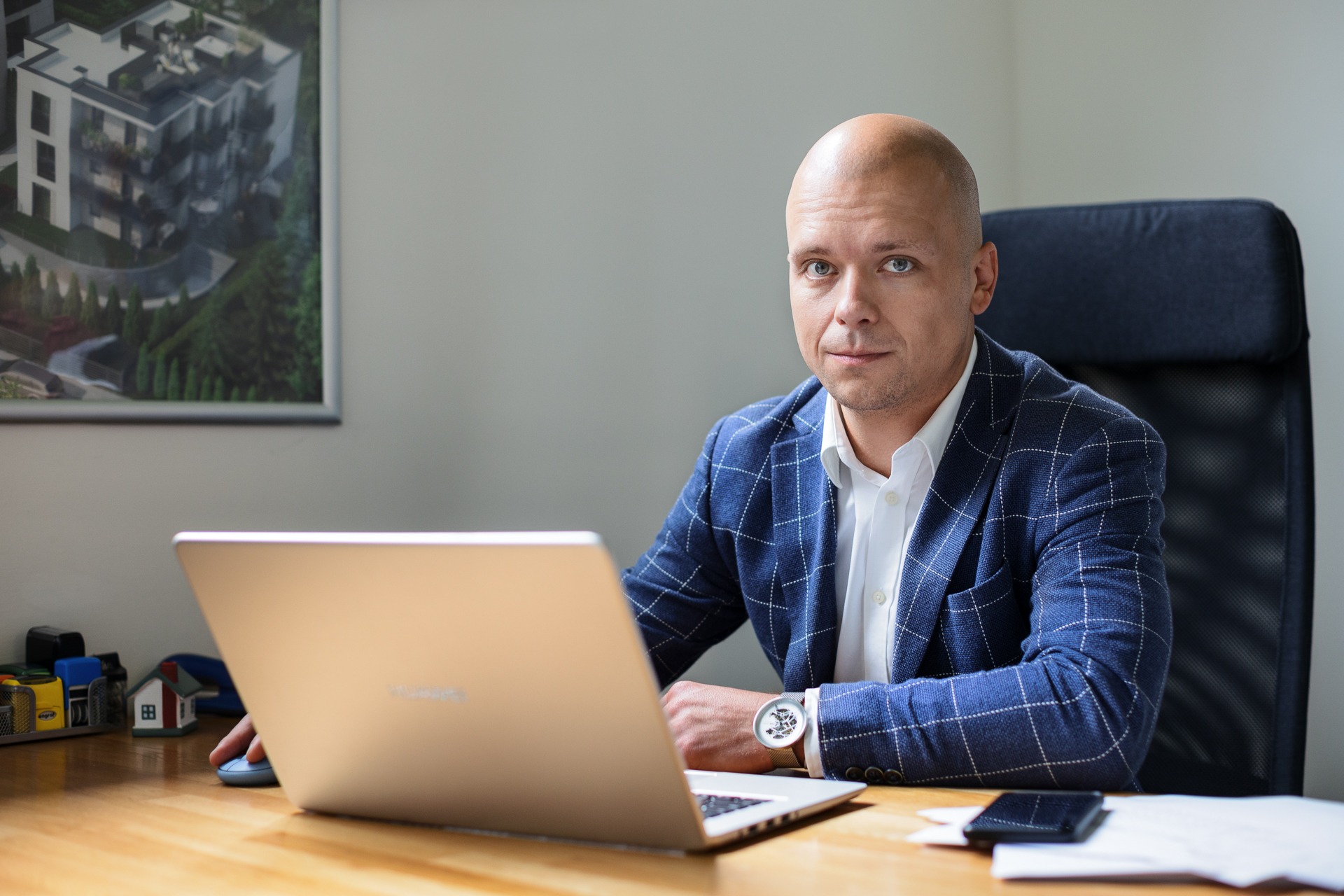 Mężczyzna w niebieskiej marynarce w kratę siedzi za biurkiem z laptopem, na ścianie wisi obraz z wizualizacją budynku mieszkalnego.