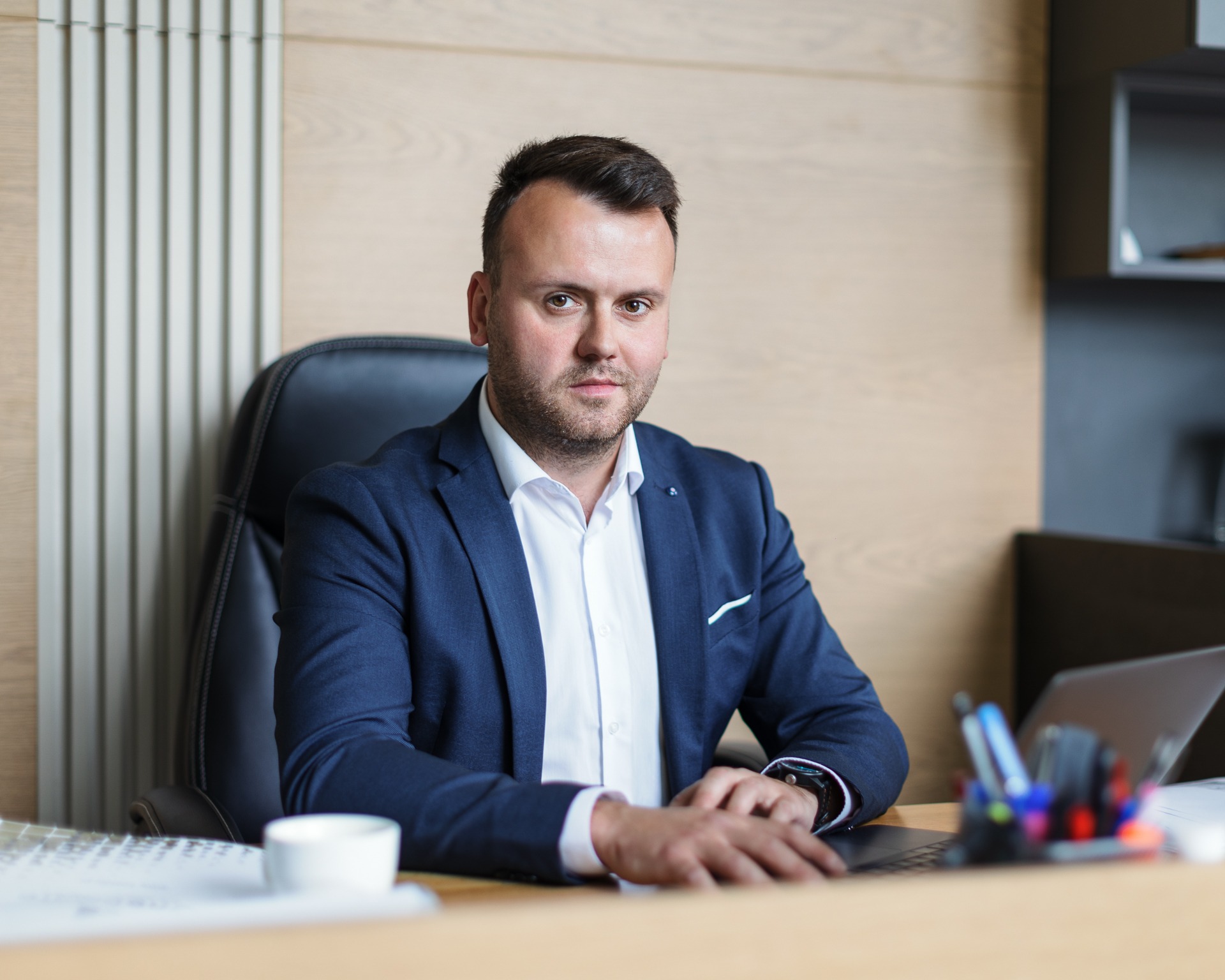 Mężczyzna w niebieskiej marynarce siedzi za biurkiem z laptopem, filiżanką i przyborami do pisania, patrząc w kamerę.