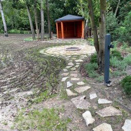 Budogrody - Ścieżka z nieregularnych kamieni prowadząca do drewnianej altany z paleniskiem na okrągłym podeście, świeżo obsiana trawa na gliniastej ziemi, widoczny słupek ogrodzeniowy.