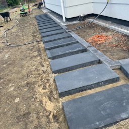 Budogrody - Układanie ścieżki z prostokątnych, grafitowych płyt betonowych wzdłuż białej elewacji domu, widoczna glebogryzarka i wózek budowlany w tle.