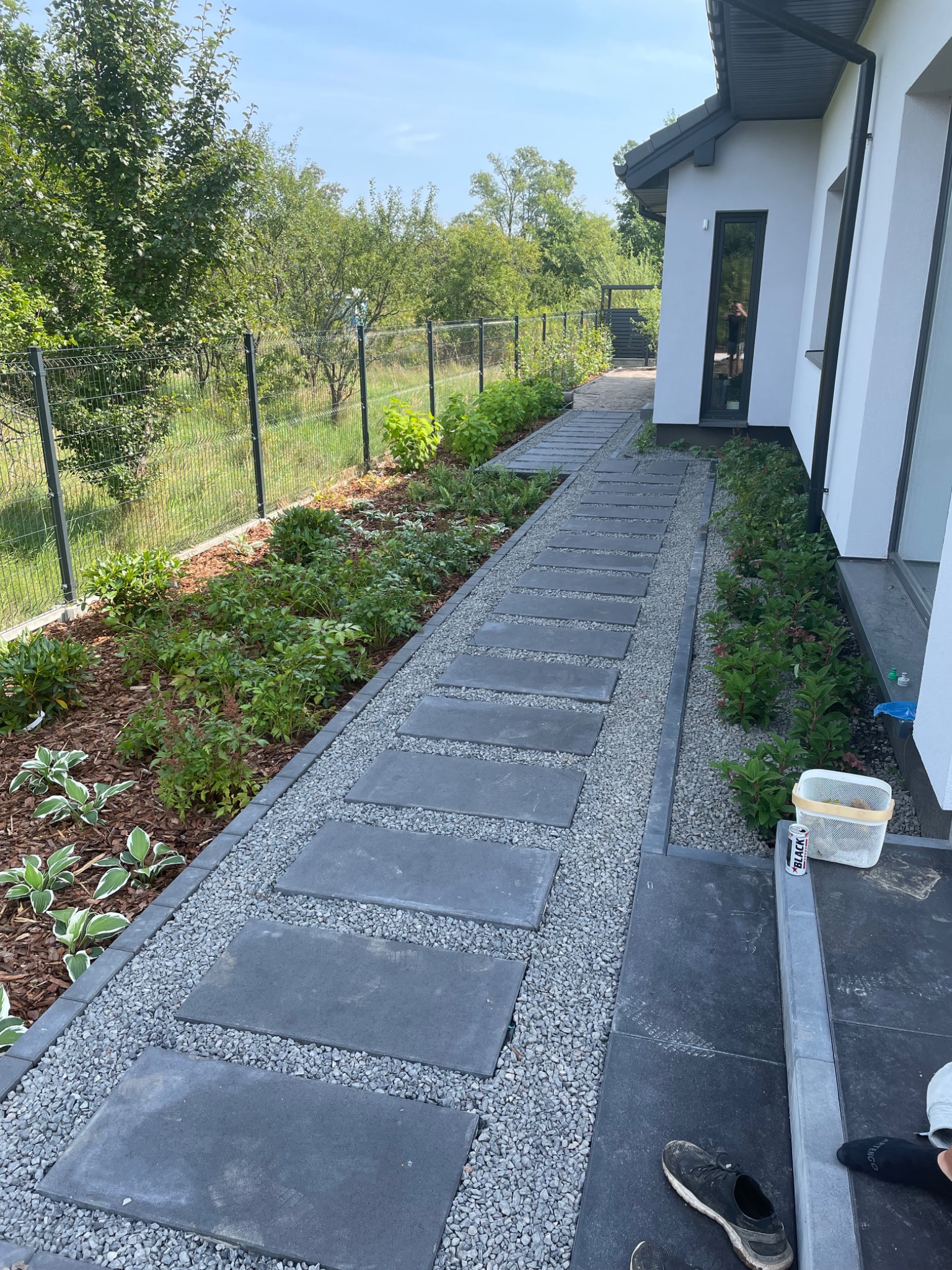 Ścieżka z prostokątnych płyt betonowych ułożonych w trawie i żwirze, prowadząca wzdłuż ściany nowoczesnego domu, z widocznym fragmentem ogrodu z roślinami i siatką ogrodzeniową.