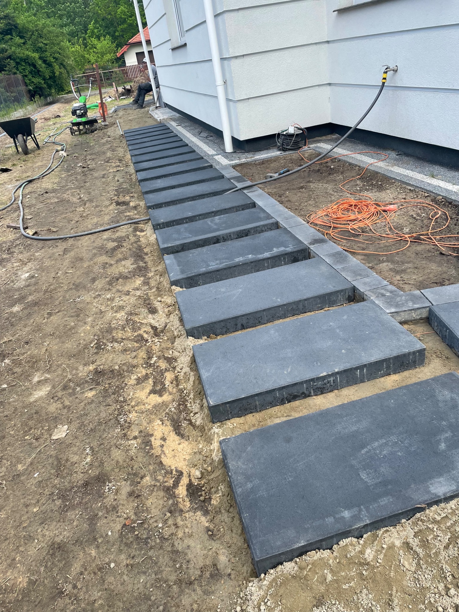 Układanie ścieżki z prostokątnych, grafitowych płyt betonowych wzdłuż białej elewacji domu, widoczna glebogryzarka i wózek budowlany w tle.