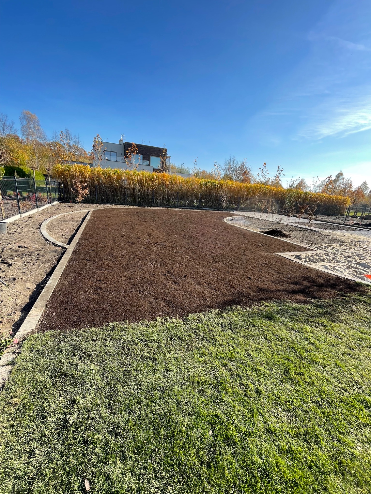 Przygotowany teren pod trawnik z rolki, obrzeża z kostki betonowej, widok na nowoczesny dom i ozdobne trawy w tle.