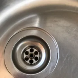 Metalowy odpływ zlewu z siedmioma otworami, widoczne zarysowania na powierzchni zlewu.