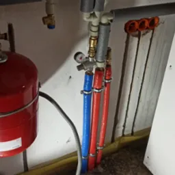 Czerwony zbiornik wyrównawczy ciśnienia obok rur instalacji centralnego ogrzewania, z przewodami w izolacji w kolorach czerwonym i niebieskim, zamocowanymi obejmami do ściany.