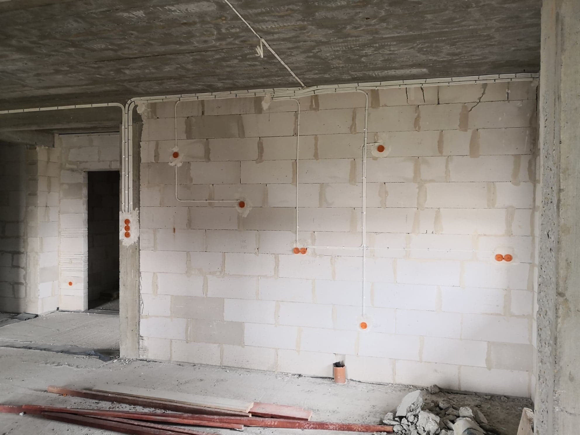 Surowy stan: białe ściany z betonu komórkowego, widoczne przewody w białych listwach i pomarańczowe puszki pod gniazdka, betonowy sufit i podłoga w szarym odcieniu, elementy drewniane na podłodze.