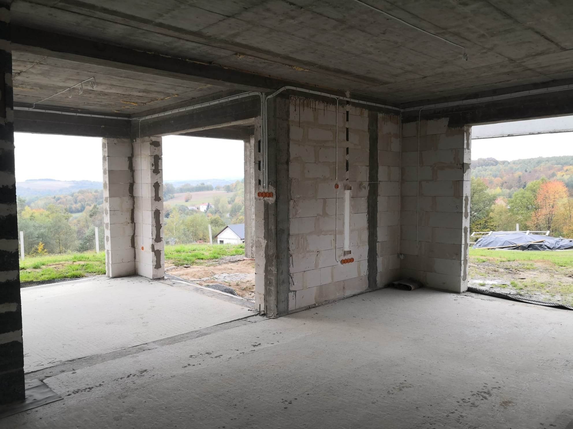 Surowy stan wnętrza budynku w trakcie budowy z widocznymi przewodami elektrycznymi prowadzonymi po ścianach i pod sufitem, puszki instalacyjne wmurowane w ściany z betonu komórkowego, widok...