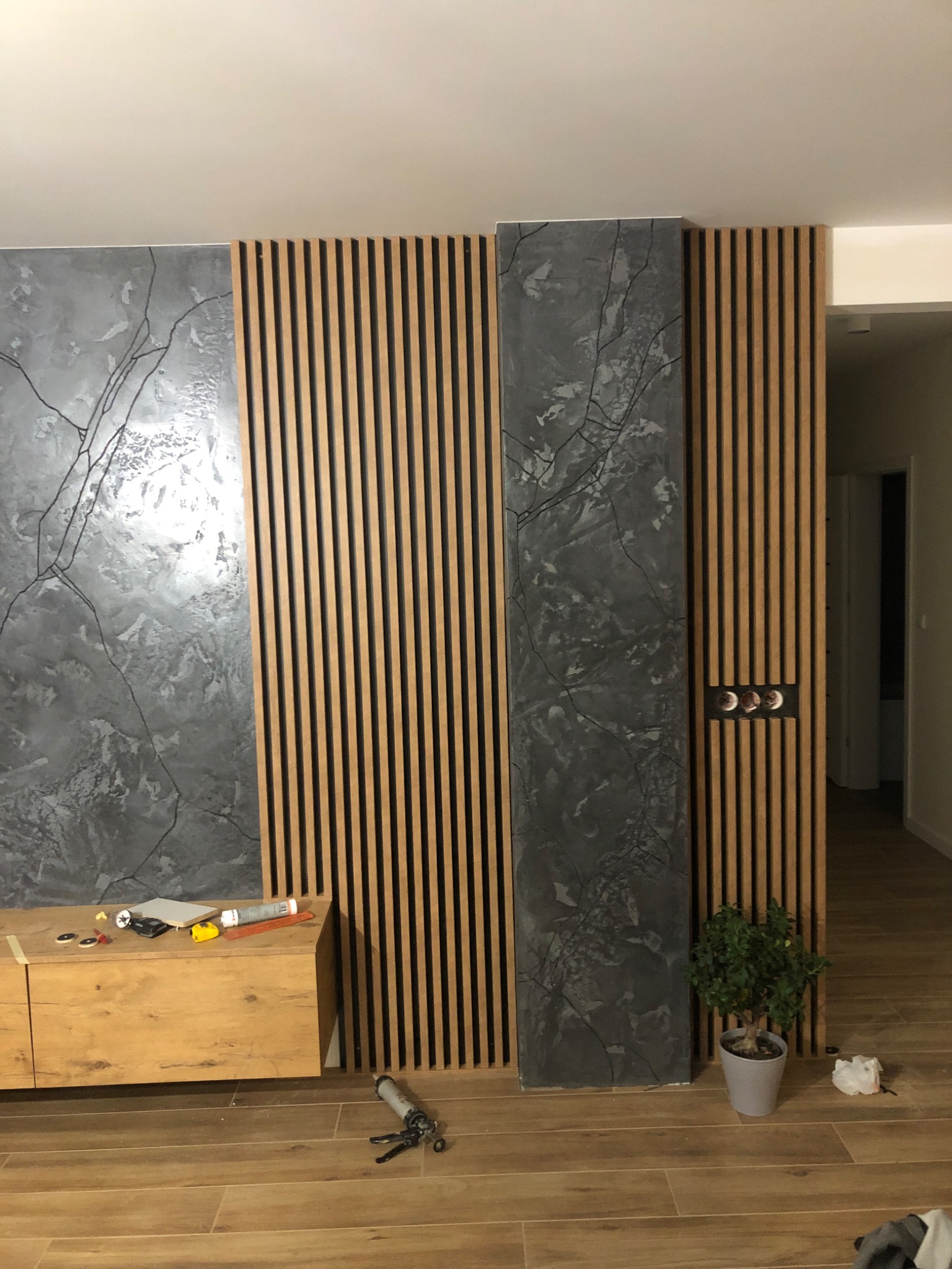 Montaż dekoracyjnej ściany z drewnianych listew i paneli imitujących marmur w salonie, widoczne narzędzia i akcesoria montażowe.