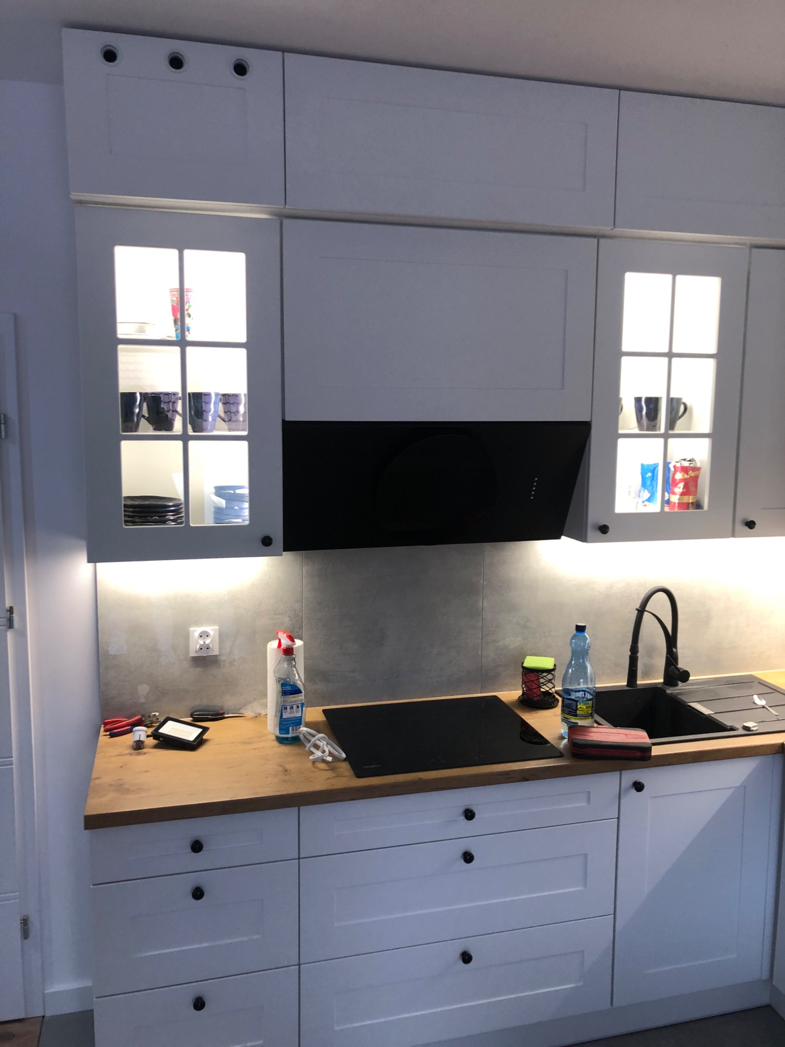 Jasna kuchnia z białymi szafkami, drewnianym blatem i czarnym okapem, oświetlenie podszafkowe, widoczne naczynia w przeszklonych szafkach wiszących.