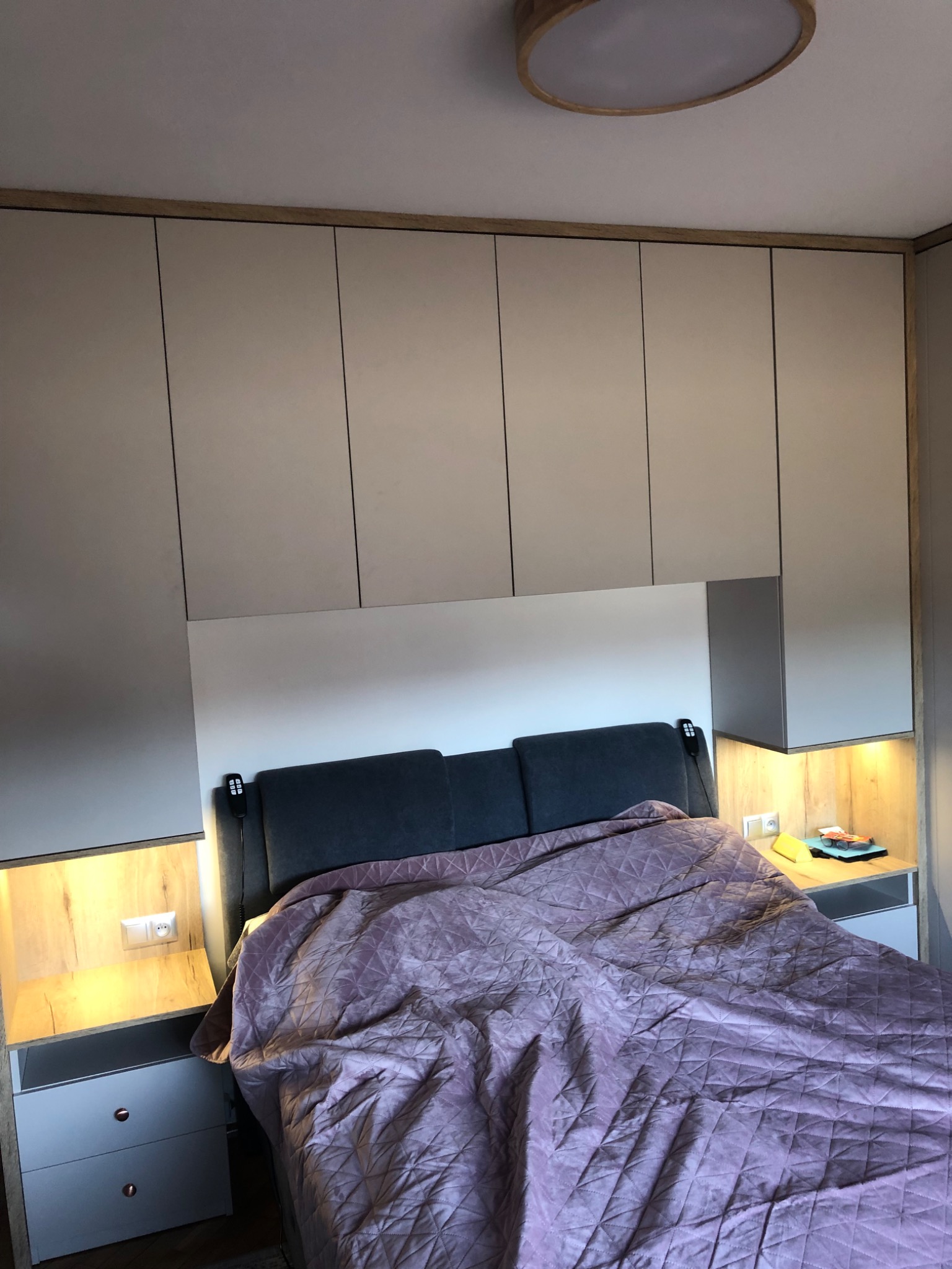 Szare szafy wiszące nad łóżkiem z zagłówkiem, oświetlone szafki nocne z gniazdkami i lampką, fioletowa narzuta na łóżku.