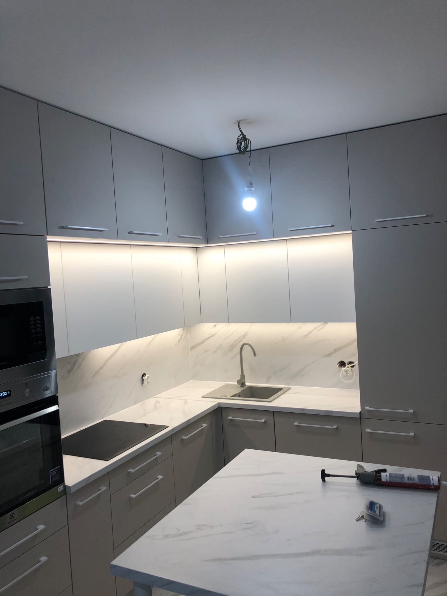 Nowoczesna kuchnia w odcieniach szarości z marmurowym blatem i podświetleniem LED, widoczne narzędzia montażowe na blacie wyspy.