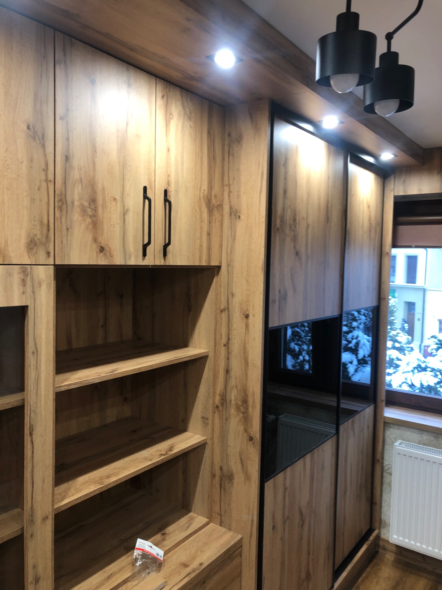 Drewniana zabudowa meblowa z szafą przesuwną z czarnymi detalami i otwartymi półkami, oświetlenie punktowe w suficie i widok za oknem na ośnieżone drzewa.