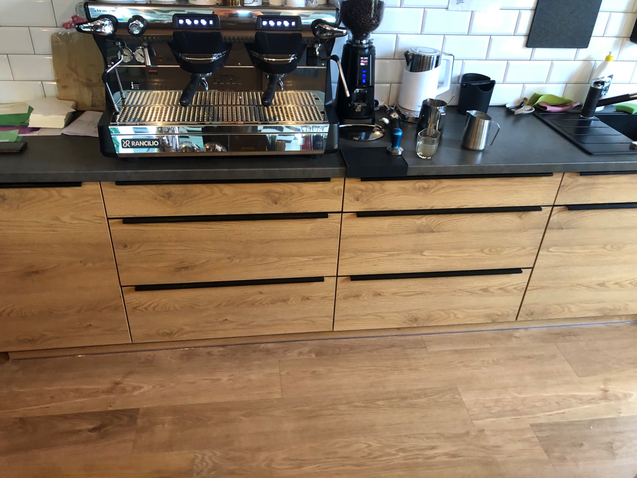 Nowoczesna zabudowa kuchenna z drewnianymi frontami i czarnymi uchwytami, na blacie ekspres do kawy i młynek, widoczna podłoga z paneli.