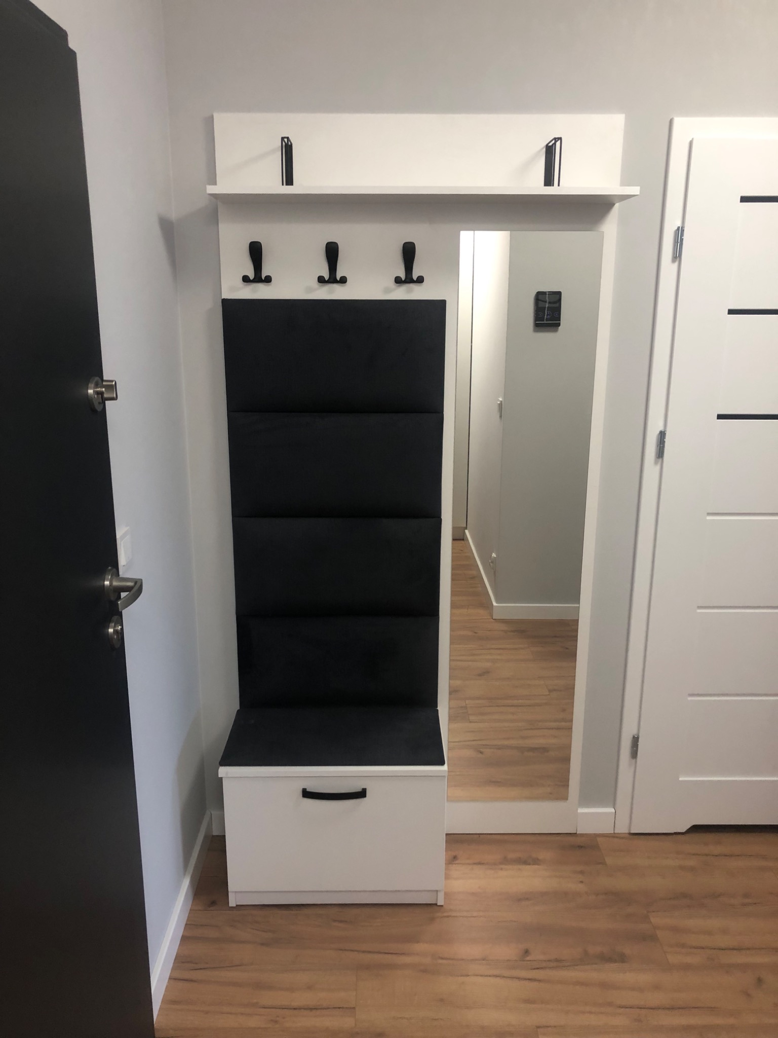 Nowoczesny przedpokój z biało-czarnym panelem ściennym z wieszakami, szafką na buty z siedziskiem i lustrem, widok z perspektywy wejścia.