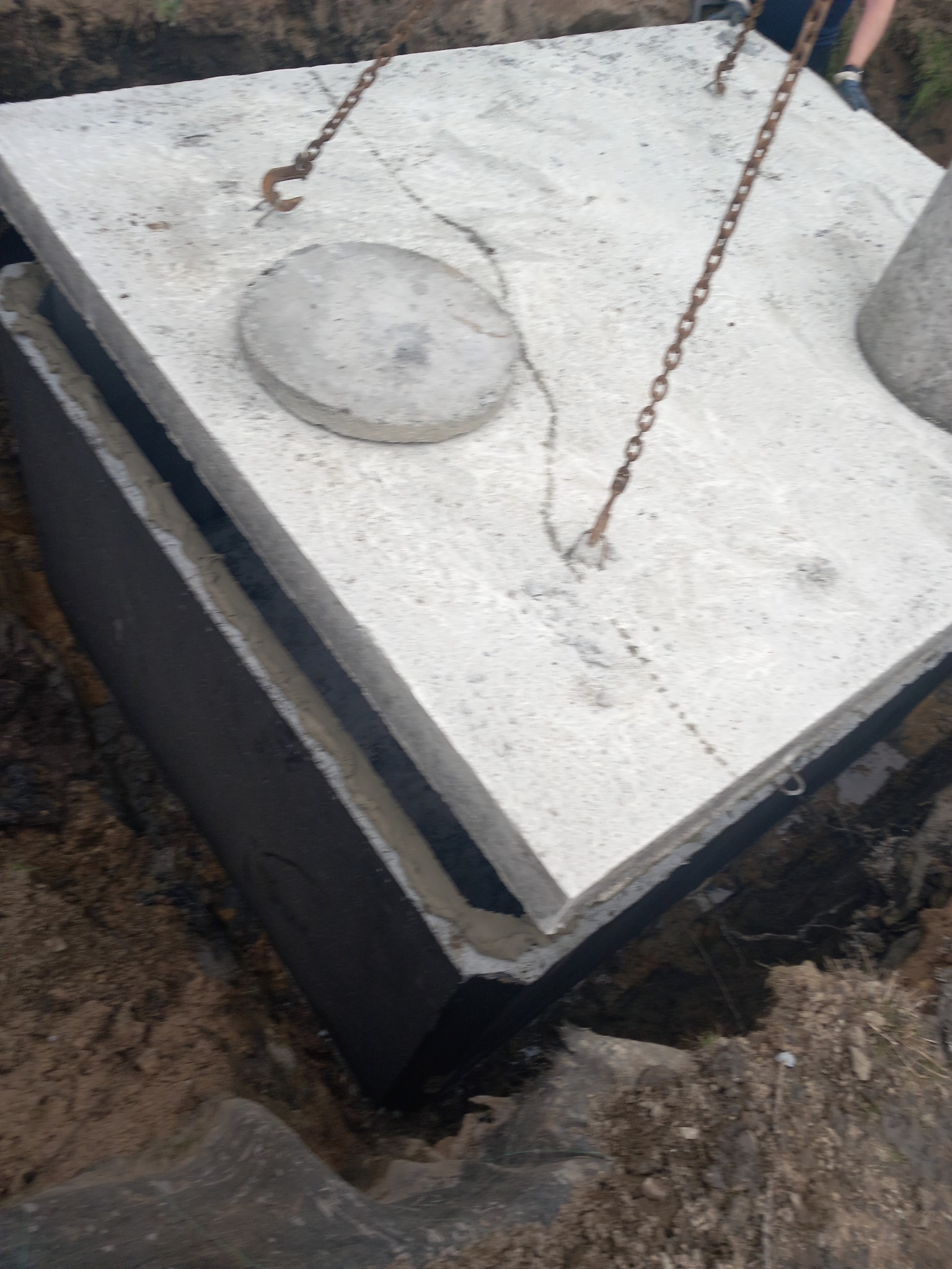 Betonowy zbiornik z pokrywą opuszczany do wykopu za pomocą łańcuchów, widoczne uszczelnienie i grunt.