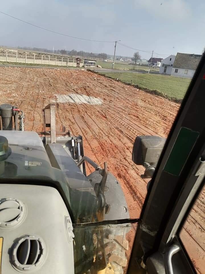 Widok z kabiny ładowarki na wyrównany teren z widocznymi śladami opon, w tle budynki gospodarcze i domy jednorodzinne.