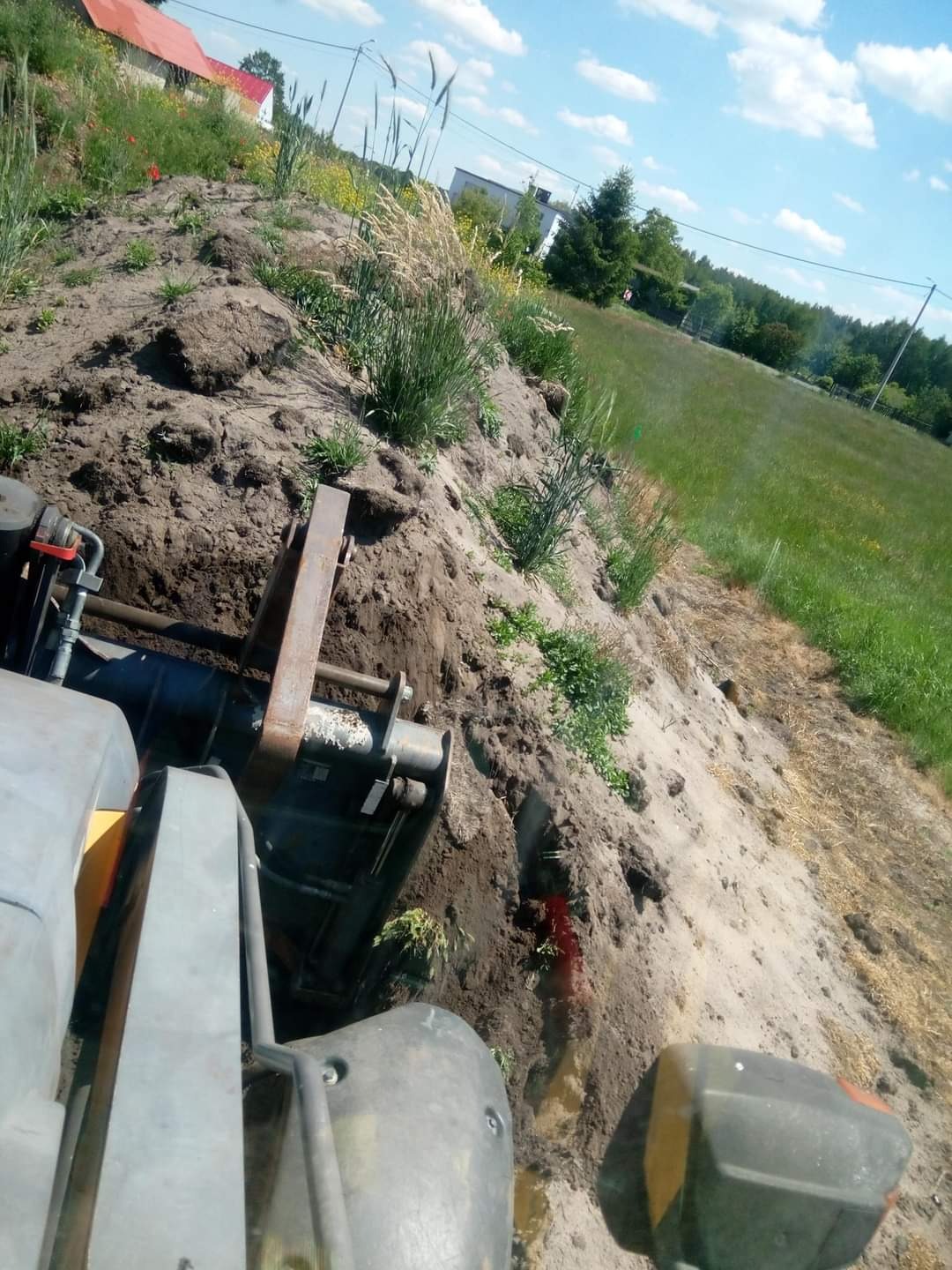 Maszyna budowlana wykonująca wykop w ziemi na tle zielonej łąki i błękitnego nieba z chmurami.