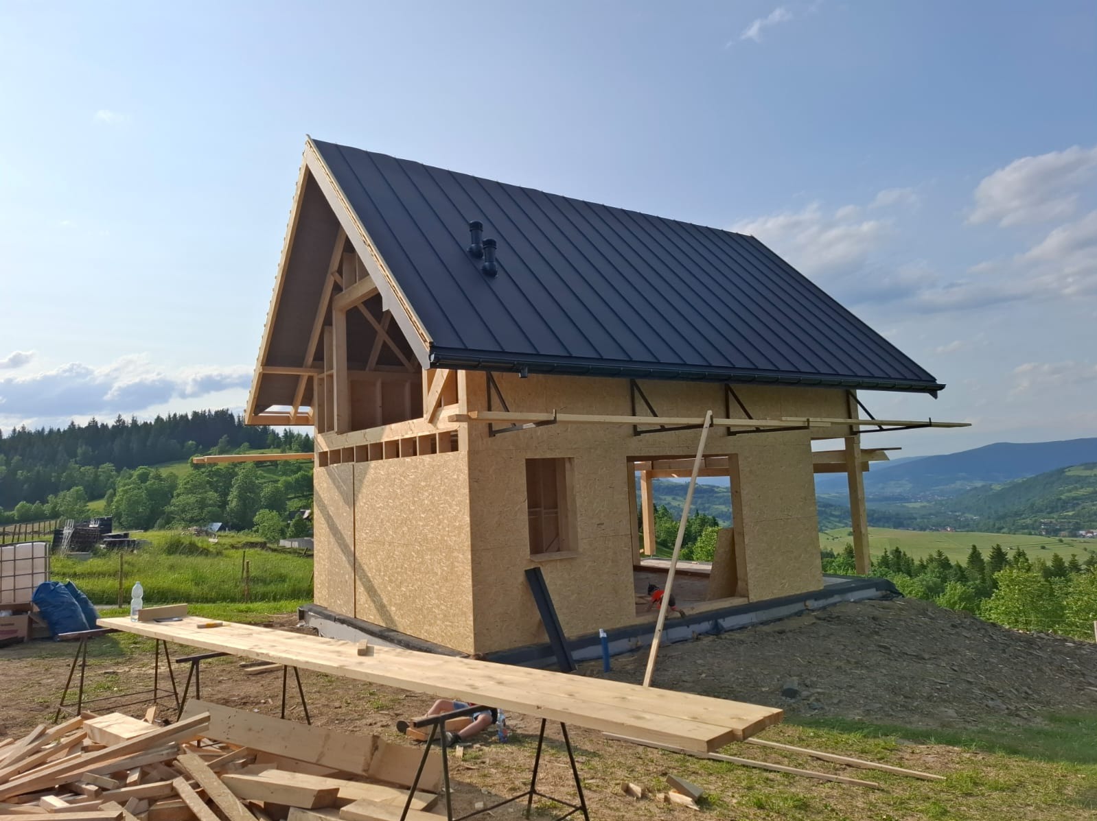 Dom szkieletowy w trakcie budowy, z widoczną konstrukcją ścian z płyt OSB i czarnym dachem, na tle zielonych wzgórz i błękitnego nieba.