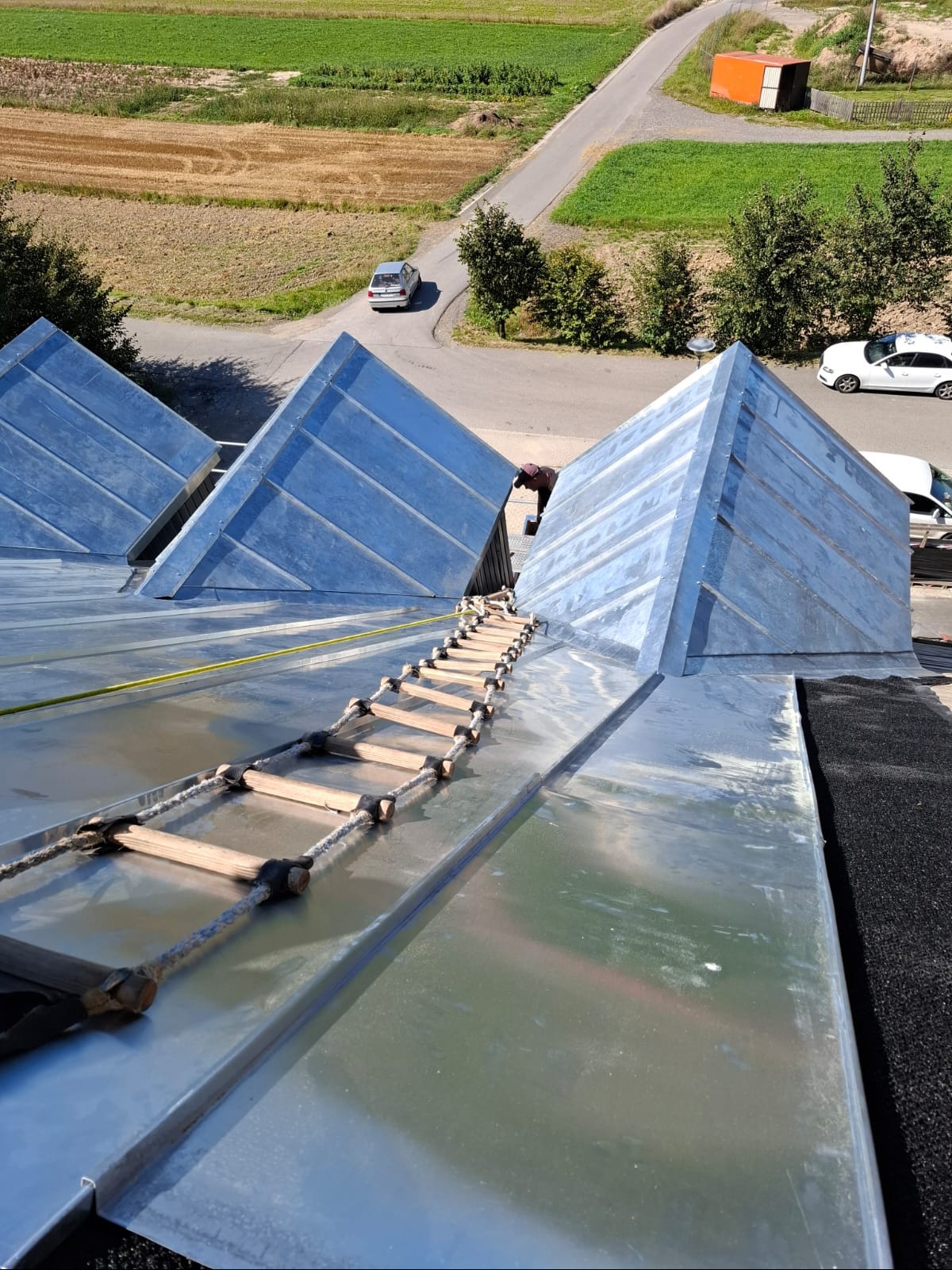 Metalowy dach z piramidalnymi lukarnami w trakcie montażu, drabina sznurowa, widok z góry na okolicę. Praca dekarza na dachu z blachy.
