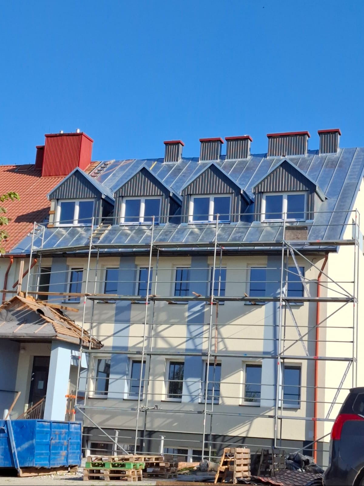 Budynek w trakcie remontu dachu z blachy, widoczne rusztowania i okna dachowe. Fragment dachu sąsiedniego budynku w kolorze czerwonym. Słoneczny dzień.