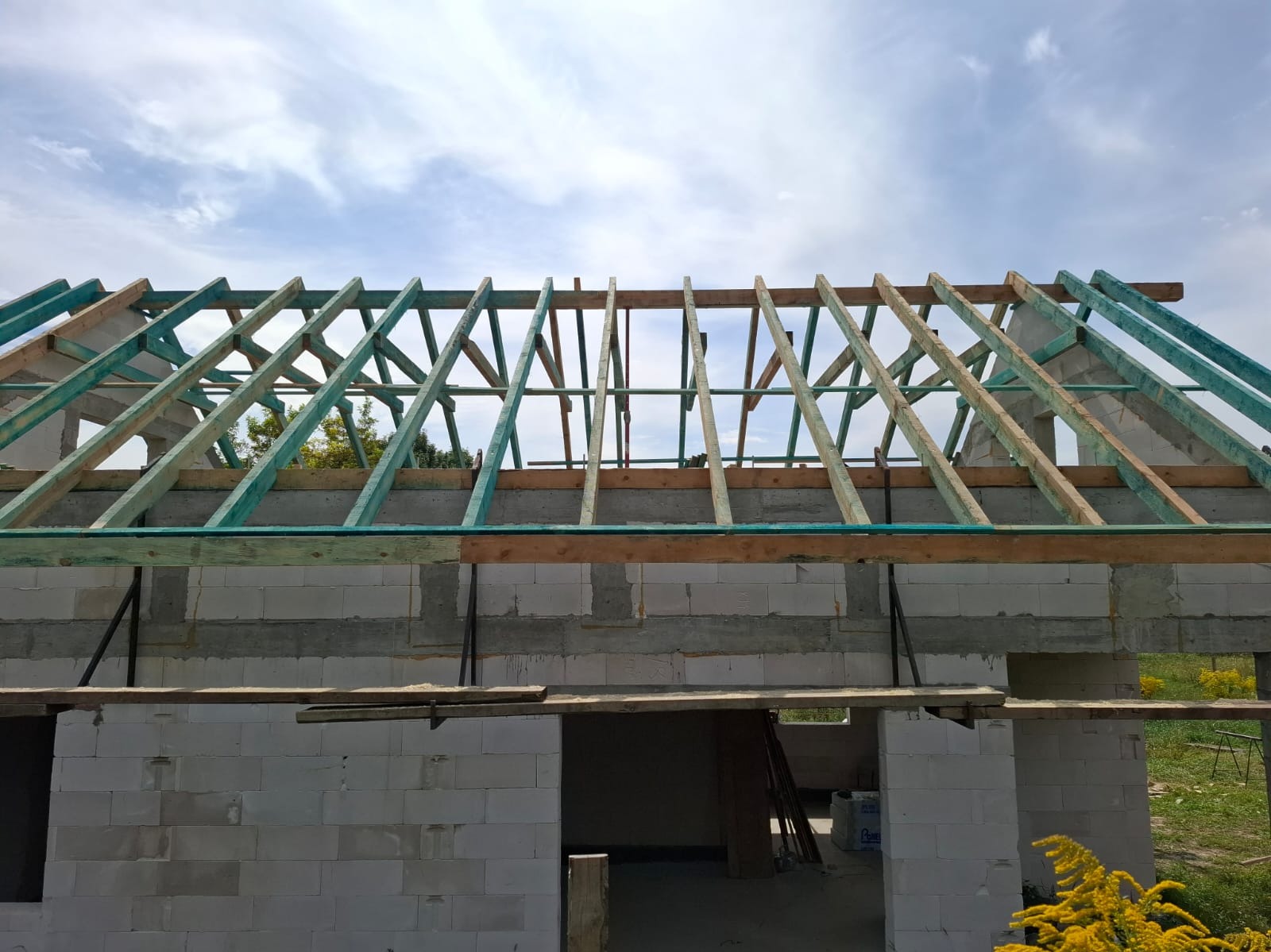 Surowy stan domu z betonu komórkowego z widoczną konstrukcją dachu z impregnowanego drewna, na tle błękitnego nieba z chmurami. Widoczne żółte kwiaty w tle.