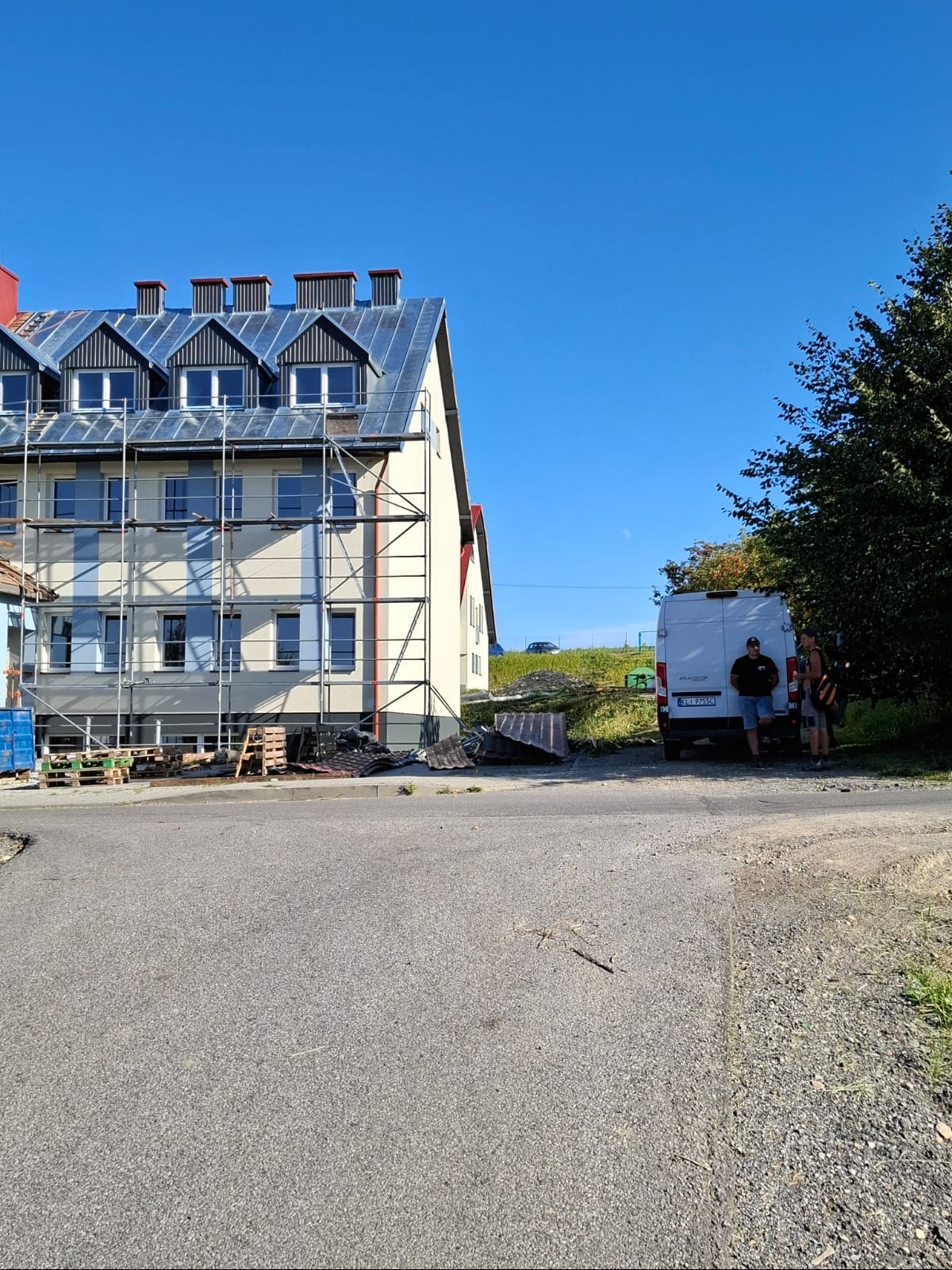 Budynek w trakcie remontu dachu, widoczne rusztowania i ekipa przy samochodzie dostawczym. Nowe pokrycie dachu z blachy. Częściowo zdjęte stare elementy.