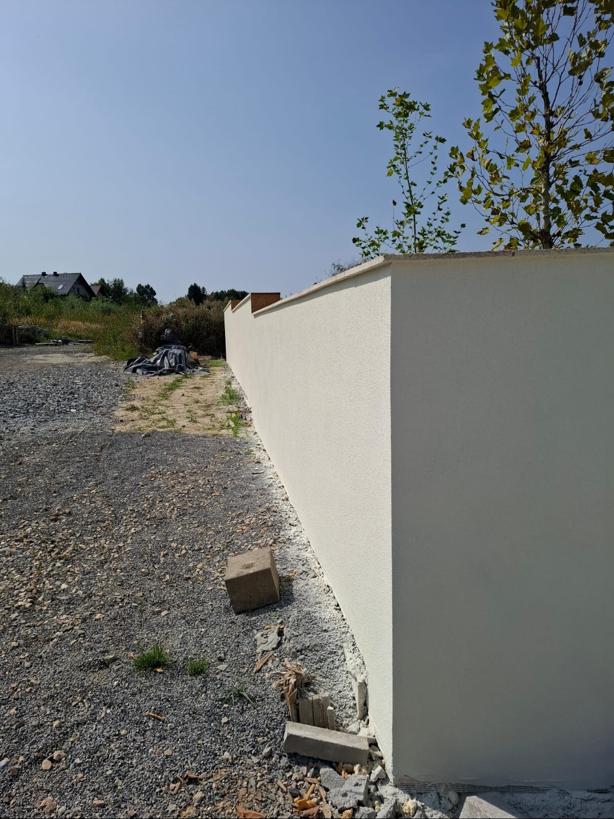 Świeżo otynkowany, biały mur ogrodzeniowy z widoczną fakturą tynku, na tle żwirowego podłoża i zieleni. Fragment drzewa w tle. Ujęcie z dołu, podkreślające solidność konstrukcji.