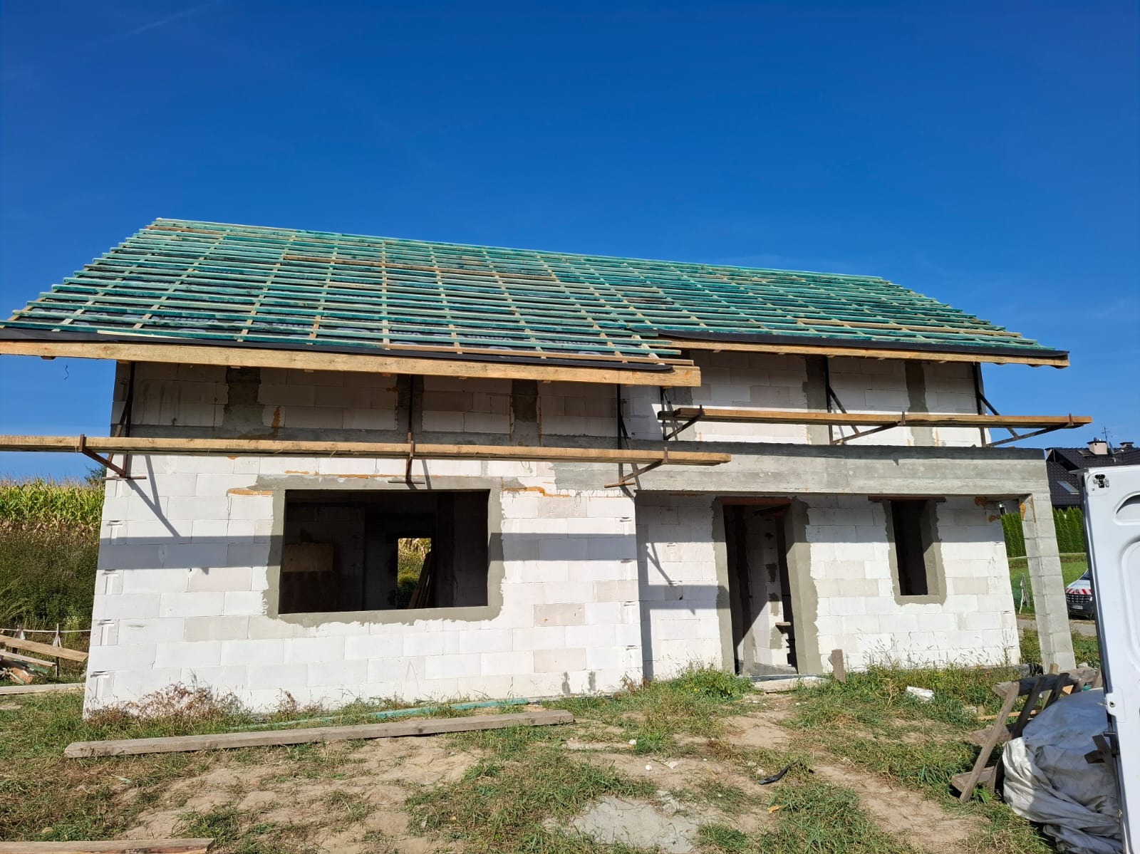 Niedokończony dom z białego pustaka z zieloną konstrukcją dachu na tle błękitnego nieba. Widoczne otwory okienne i drzwiowe, drewniane elementy konstrukcyjne.