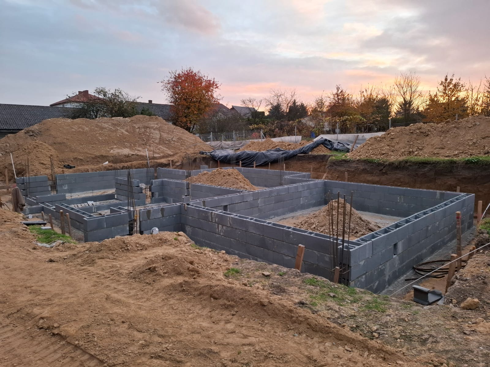 Wykopane fundamenty z bloczków betonowych, częściowo wypełnione ziemią i zbrojeniem, na tle zachodzącego słońca i jesiennych drzew.