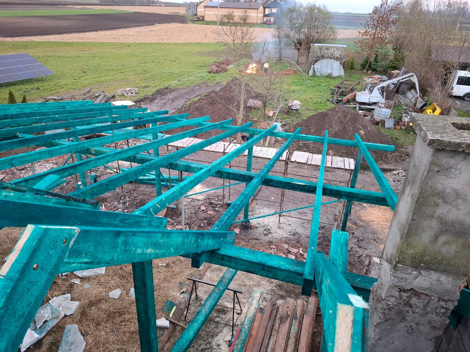 Konstrukcja dachu z impregnowanego drewna w kolorze turkusowym, widok z góry na tle wiejskiego krajobrazu z paleniskiem i zabudowaniami.