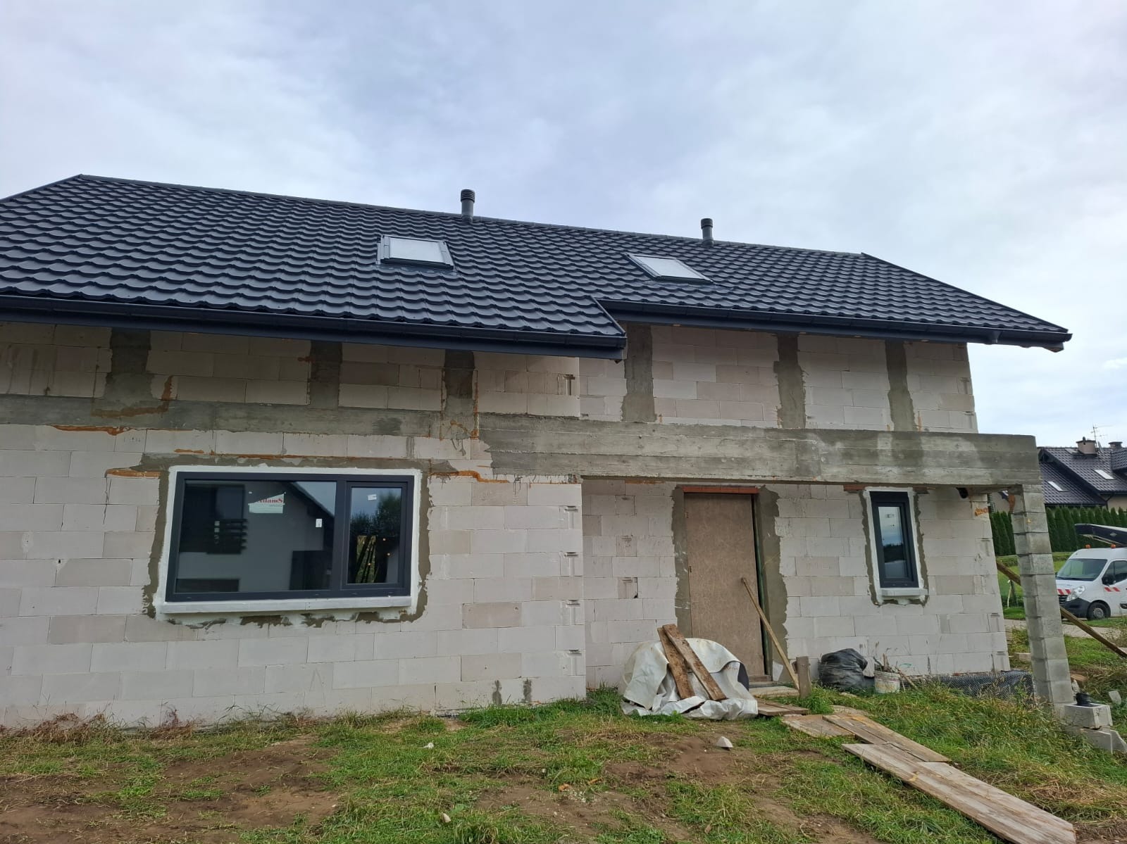 Niedokończony dom z białego bloczka z czarnym dachem z dachówki, widoczne okna i przygotowane otwory, elementy konstrukcyjne, teren budowy.