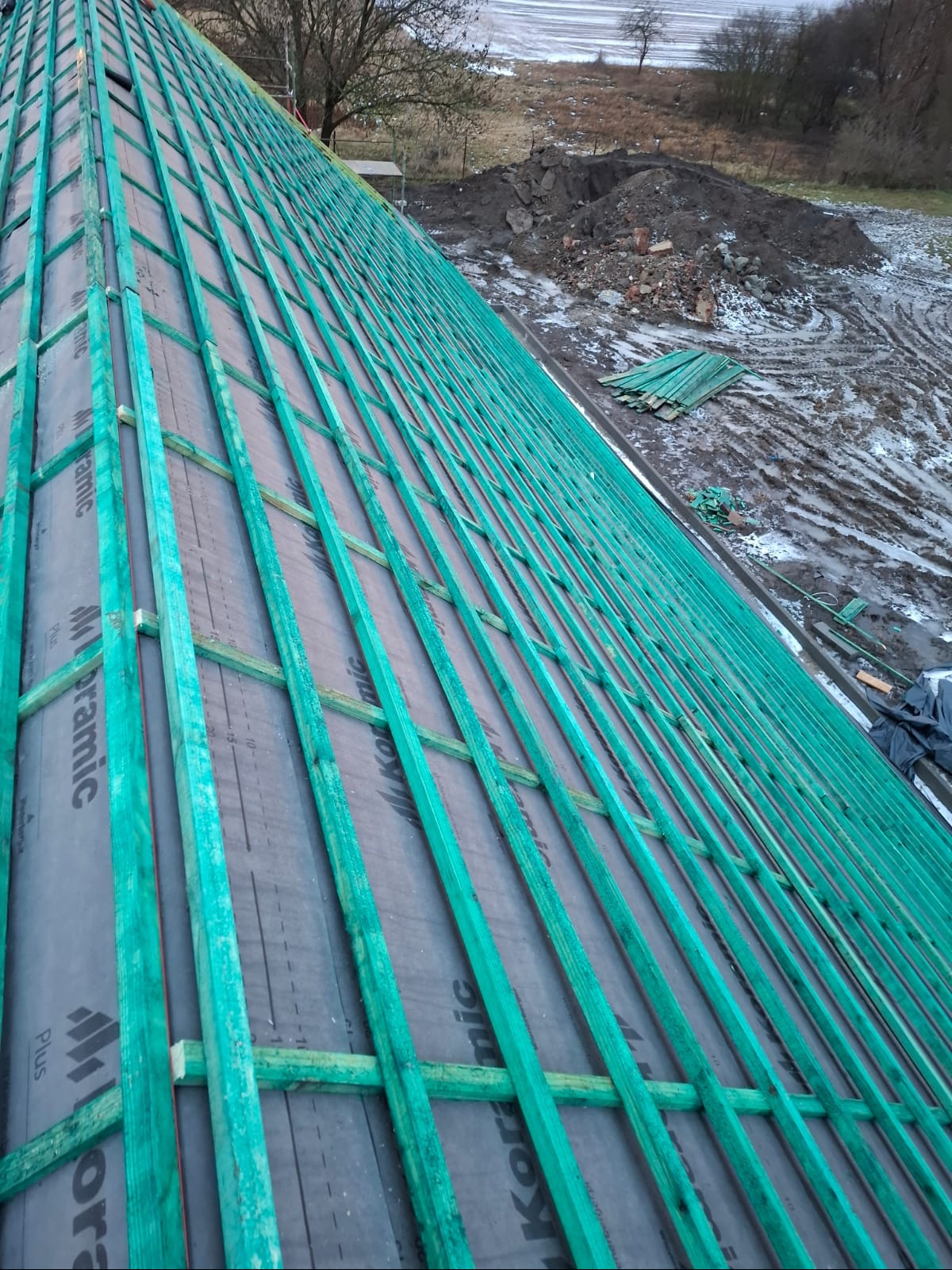 Konstrukcja dachu z zielonymi łatami na czarnej membranie, widok z góry. W tle ziemia i resztki materiałów budowlanych.