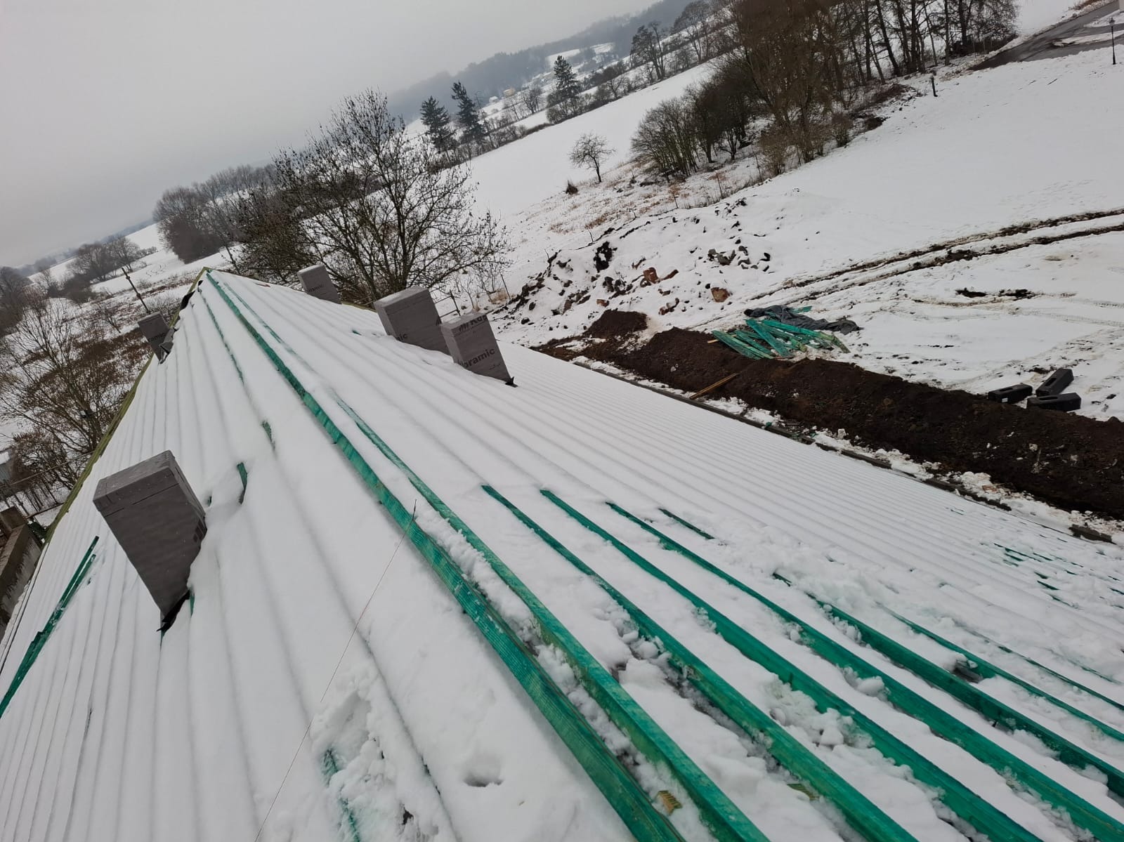 Śnieżny dach w trakcie budowy z widocznymi łatami i pustakami wentylacyjnymi BRAMIC, zimowy krajobraz w tle. Ujęcie z góry, pochmurny dzień.