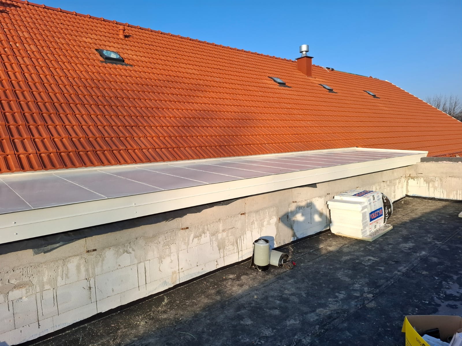 Czerwony dach z dachówkami i oknami dachowymi, widok z płaskiego dachu z izolacją i elementami instalacji. Błękitne niebo w tle.