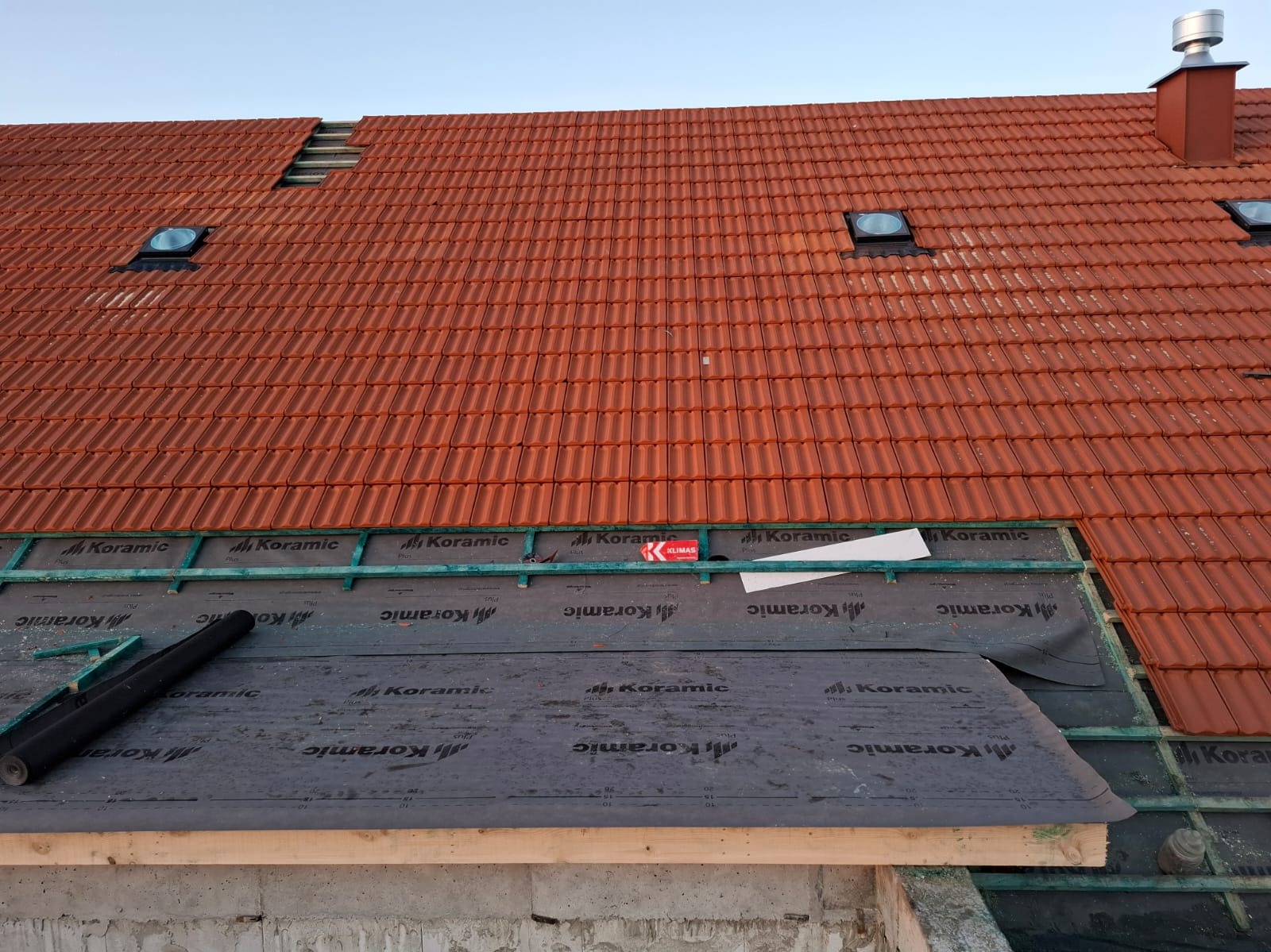 Częściowo pokryty dach z czerwoną dachówką ceramiczną Koramic, widoczne okna dachowe i komin. Fragment dachu z membraną i łatami, przygotowany do dalszego krycia.