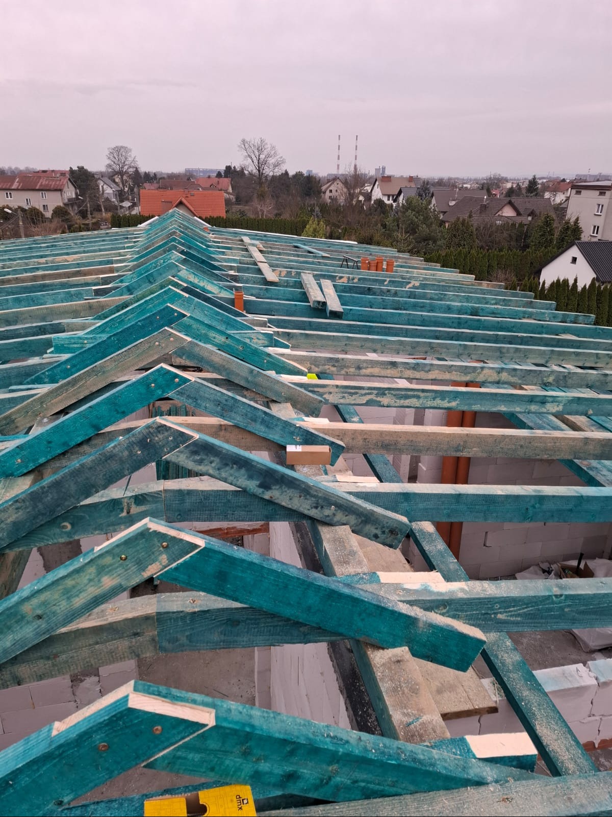 Konstrukcja dachu z impregnowanego drewna w kolorze turkusowym, widok z góry na tle zabudowy miejskiej i pochmurnego nieba. Etap budowy więźby dachowej.